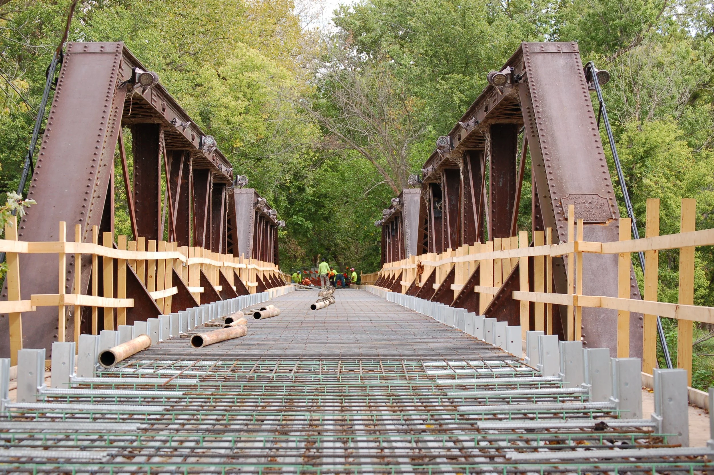 Under construction bridge with workers in the distance, wooden and metal railings, pipes on the deck, surrounded by green trees.