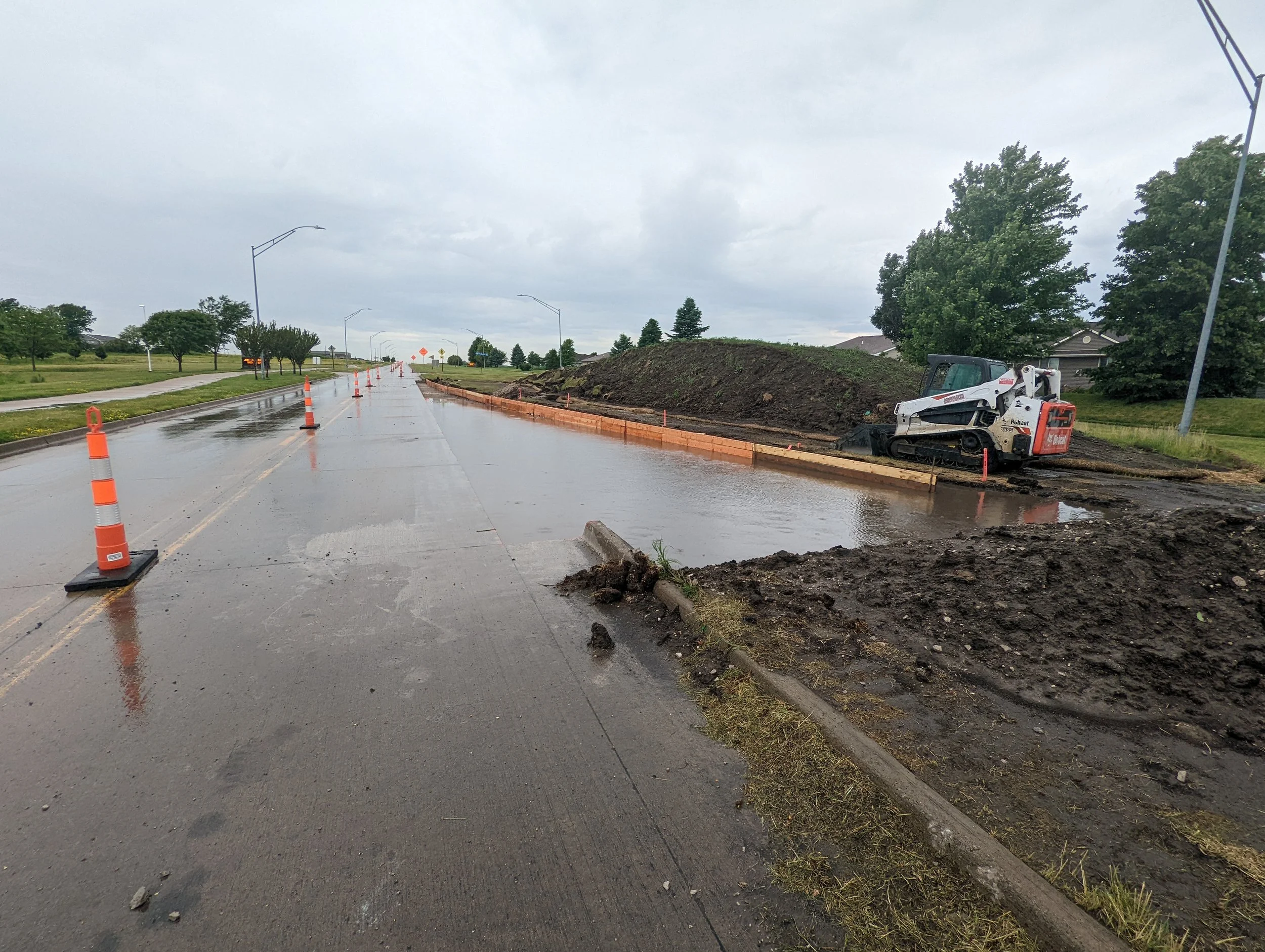 Road construction site on a rainy day with orange traffic cones, a small excavator, and a muddy sidewalk, with trees and a cloudy sky in the background.