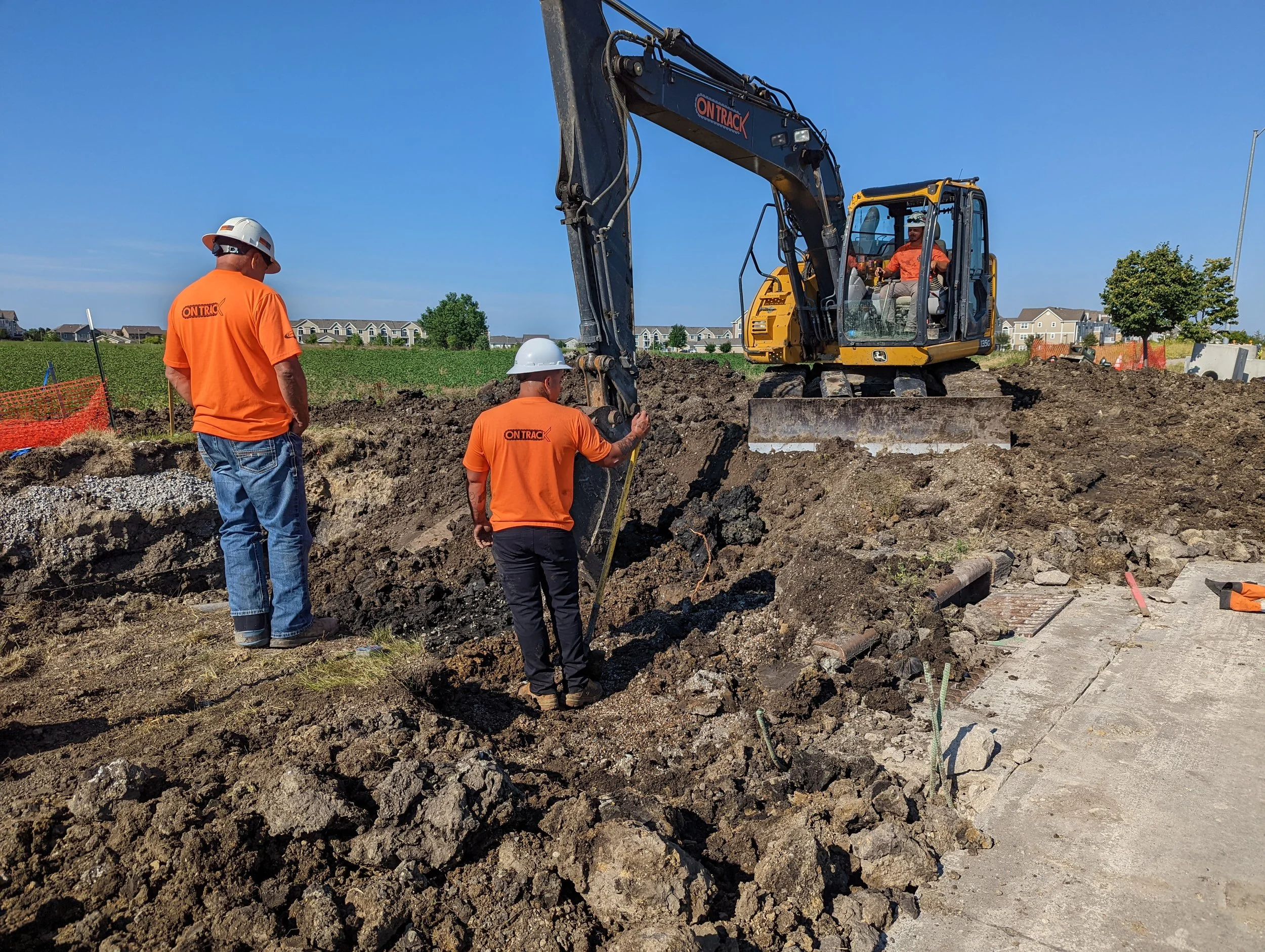 Construction workers operating a small excavator on an outdoor construction site, digging into the ground with some workers standing nearby and observing.