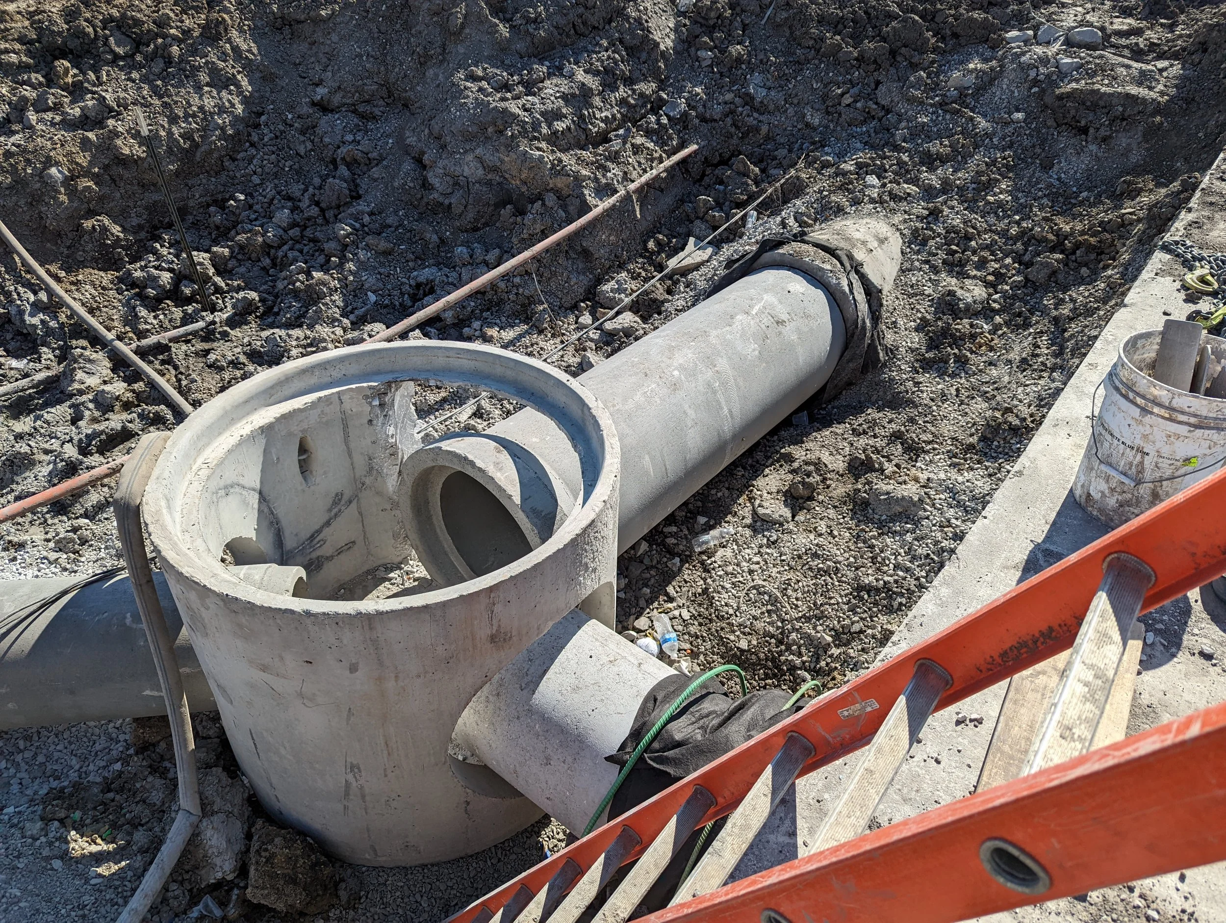 Construction site with a large concrete pipe and a specialized fitting for connecting the pipe sections, surrounded by dirt, construction tools, and safety equipment.