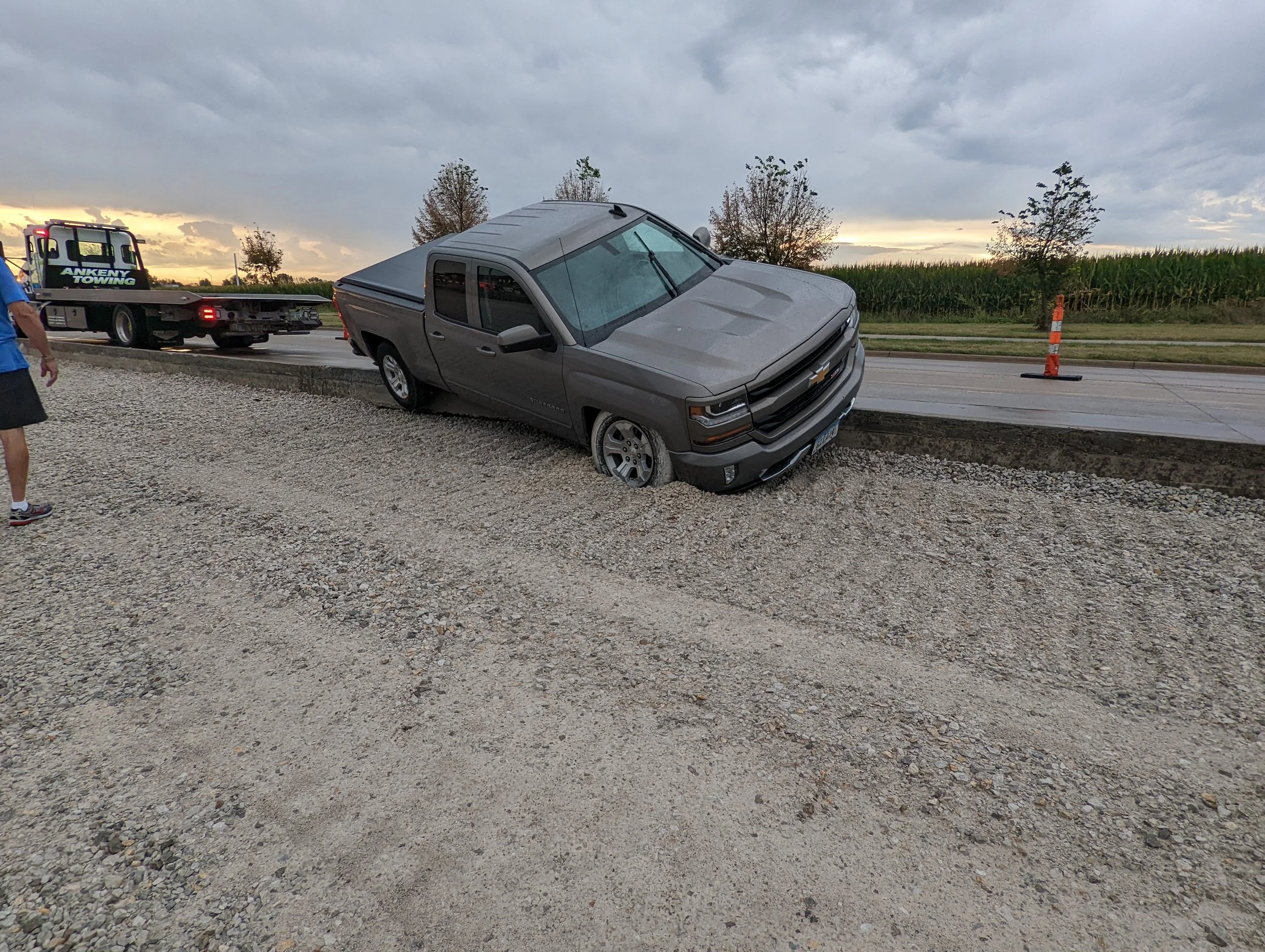 A gray pickup truck has veered off the road and is partially sunk into a gravel ditch. The truck's front end is slightly tilted downward. A tow truck is approaching from the left. There is a person standing on the gravel near the truck, and traffic c