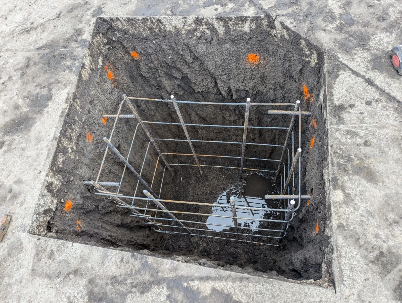 Square hole in the ground with metal rebar reinforcement and some water at the bottom, surrounded by gray concrete pavement. Orange markers are on the edges of the hole.