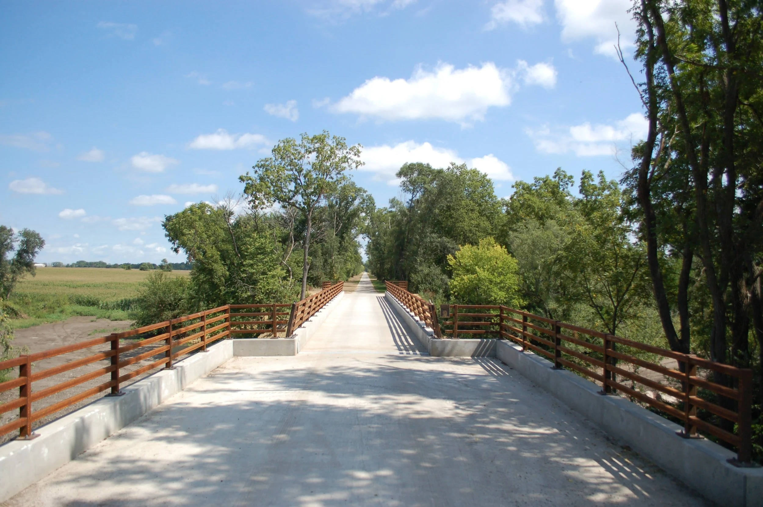 A concrete bridge with steel railings extends over a rural landscape, surrounded by green trees under a partly cloudy sky.