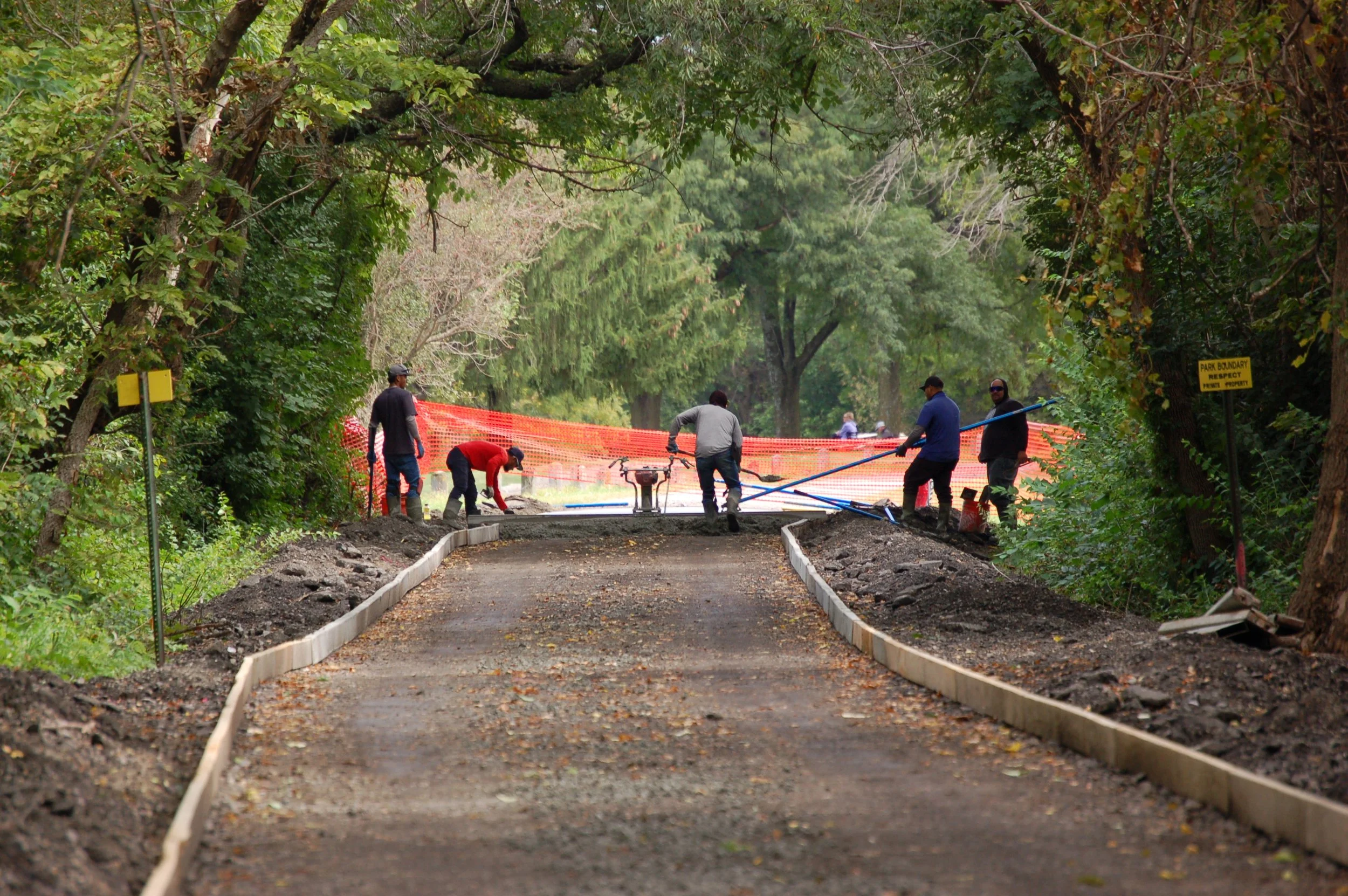 Construction workers are laying a new gravel pathway in a park, surrounded by green trees and orange safety fencing.