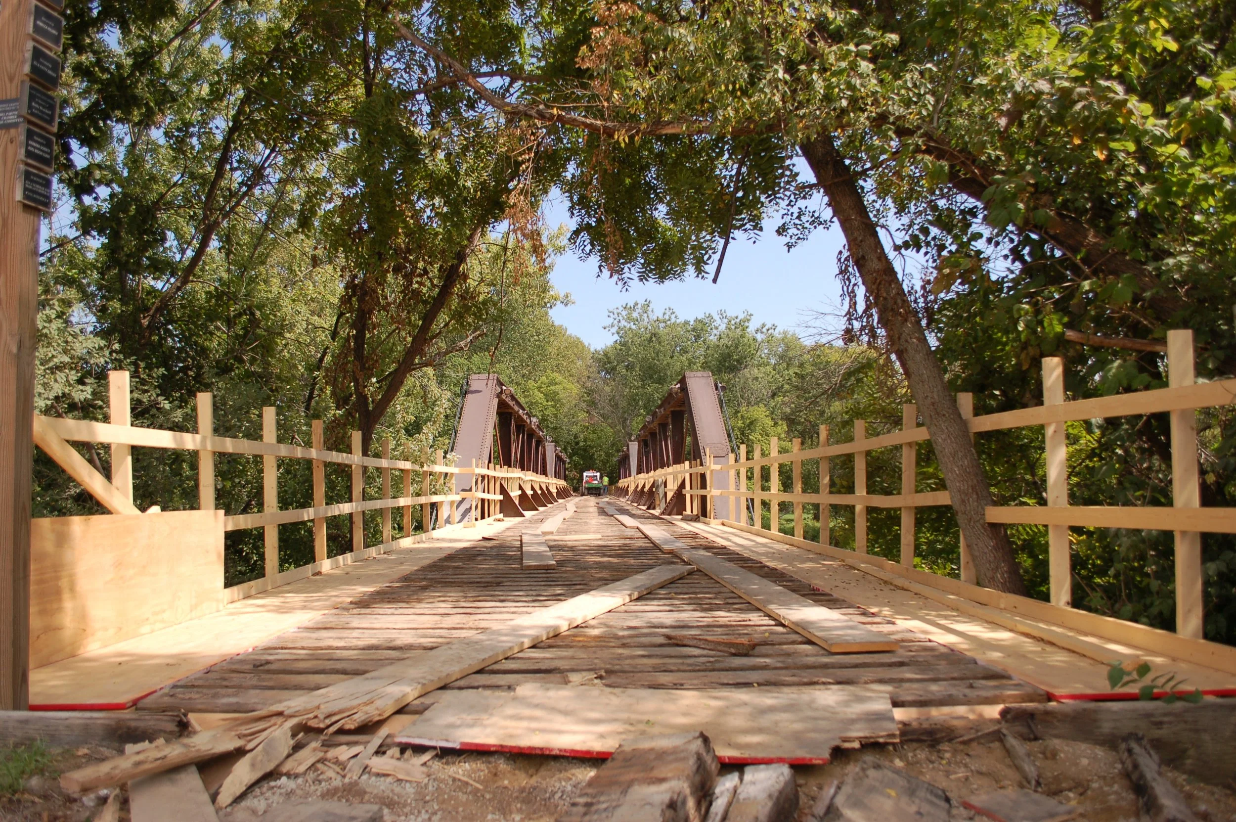 Construction of a wooden bridge being built over a small creek, surrounded by green trees, with a vehicle and construction equipment visible in the distance.