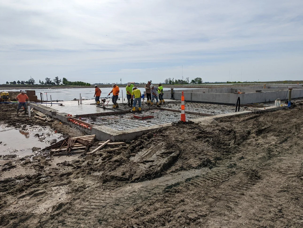 Construction workers pouring concrete on a wastewater treatment facility foundation outdoors with construction equipment and orange traffic cones nearby.