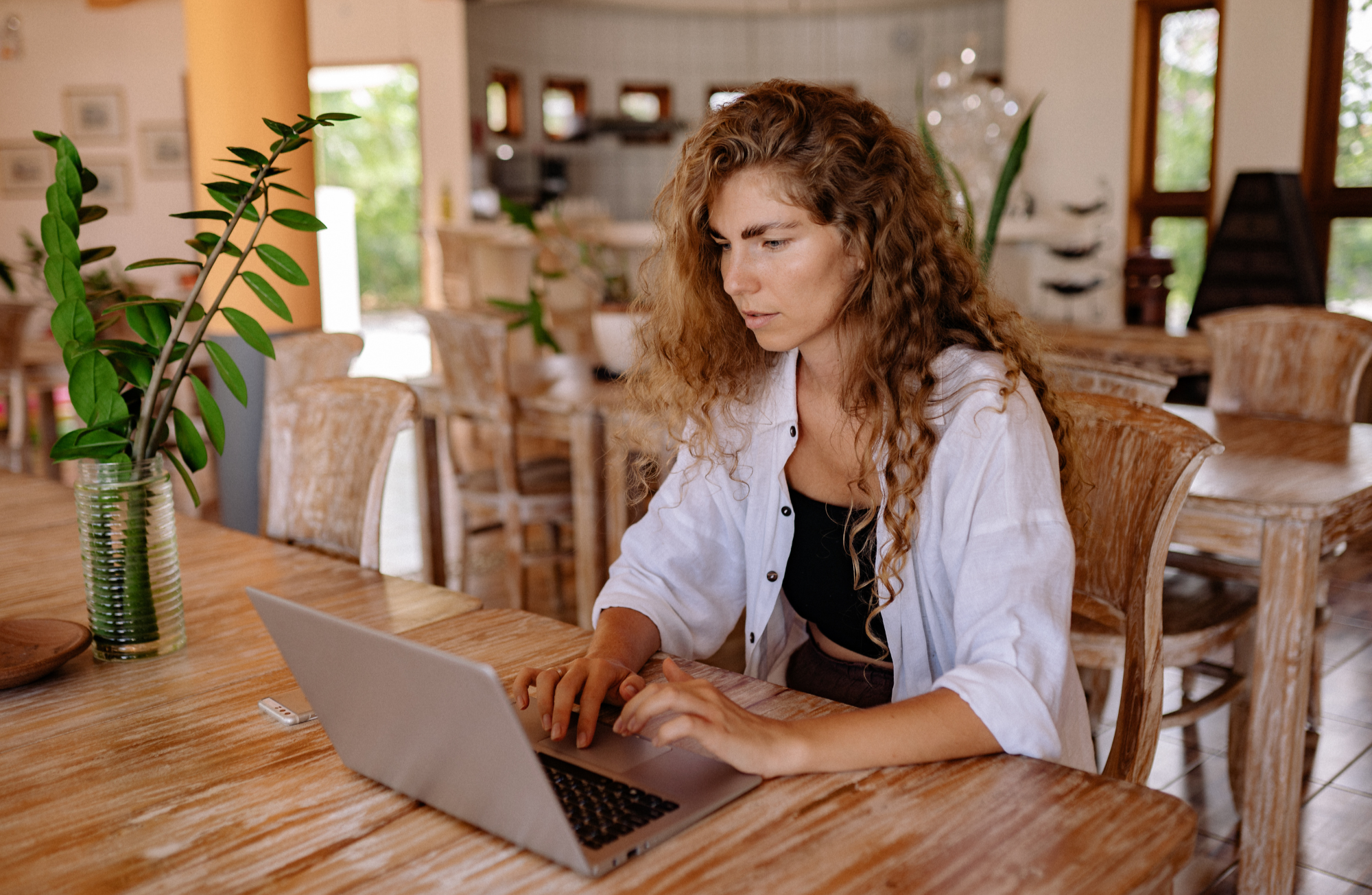 A woman with curly red hair working on a silver laptop at a wooden dining table in a well-lit, cozy home environment. Green plants and large windows are visible in the background.