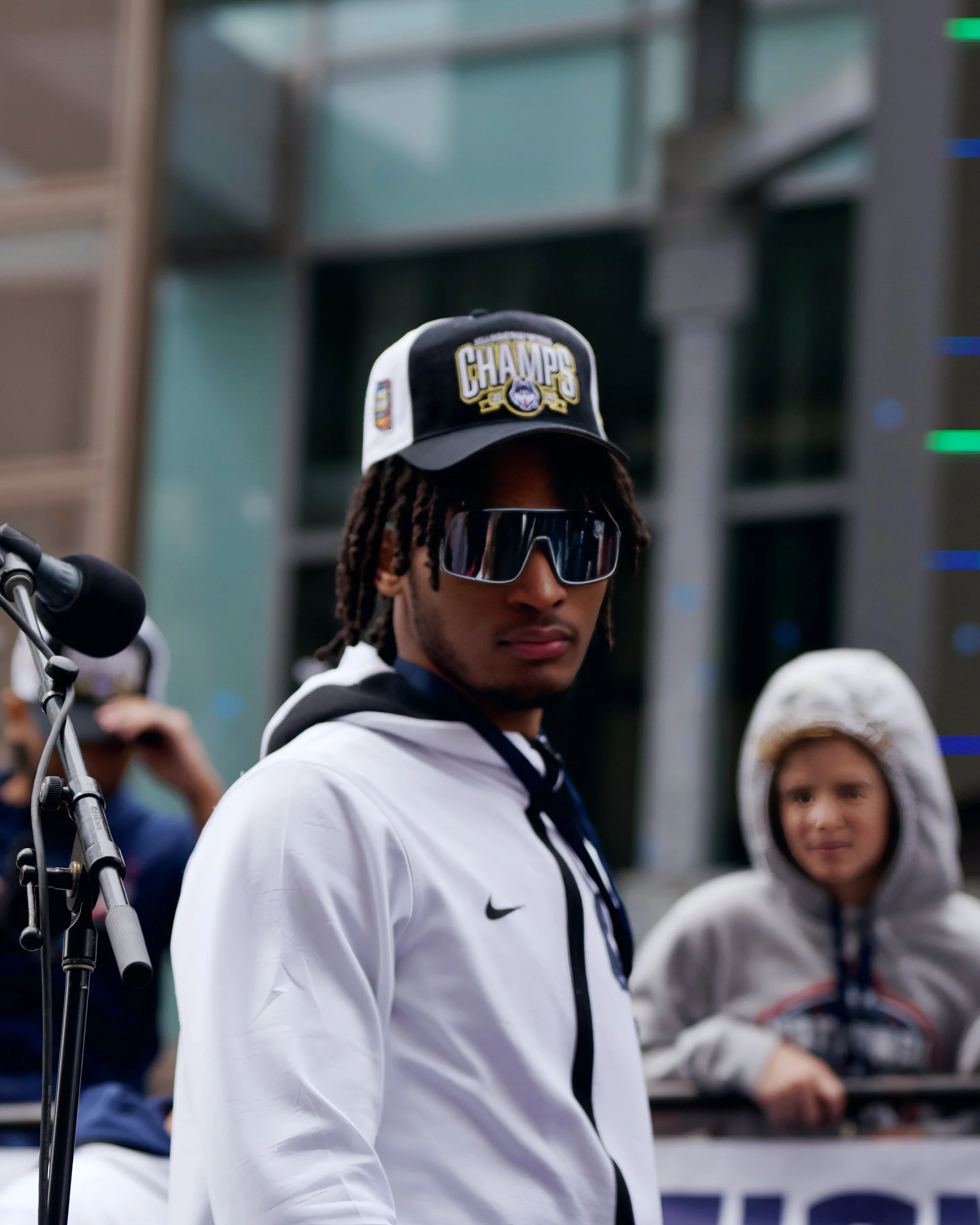 A person wearing a black and white cap with the word 'CHAMPS' embroidered on it and sunglasses, surrounded by a group of people, in an indoor setting.