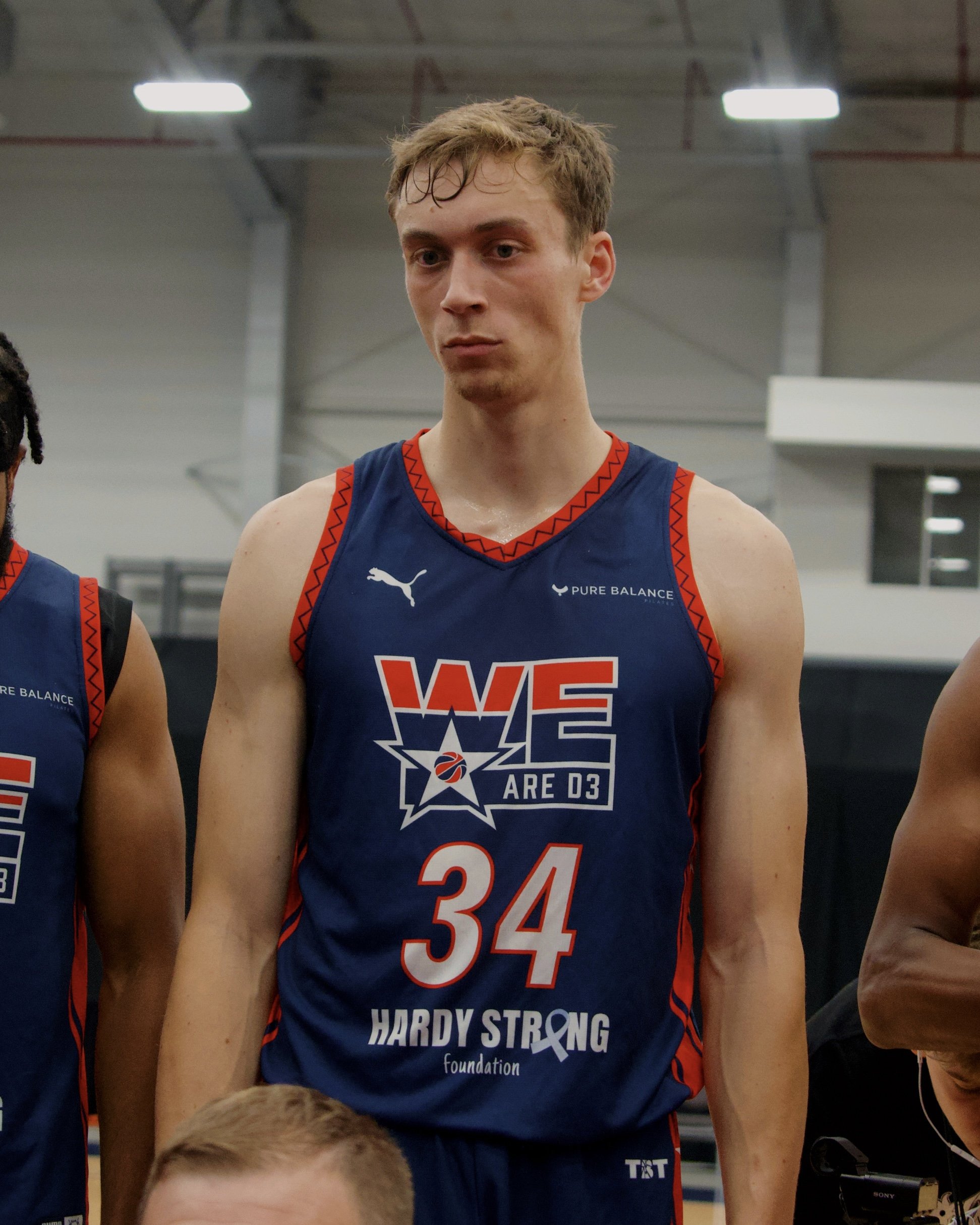 A young male basketball player wearing a navy blue jersey with red trim, featuring the number 34 and the text "HARDY STRONG foundation." He has light brown hair, a serious expression, and is standing indoors under bright lighting.