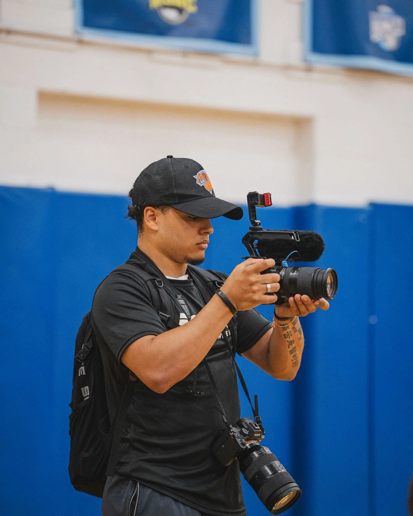 A man wearing a black Brooklyn Nets cap and black T-shirt, holding a professional camera with a large telephoto lens. He is standing in front of a blue wall, with a small camera hanging from his neck, and another camera hanging from his shoulder.