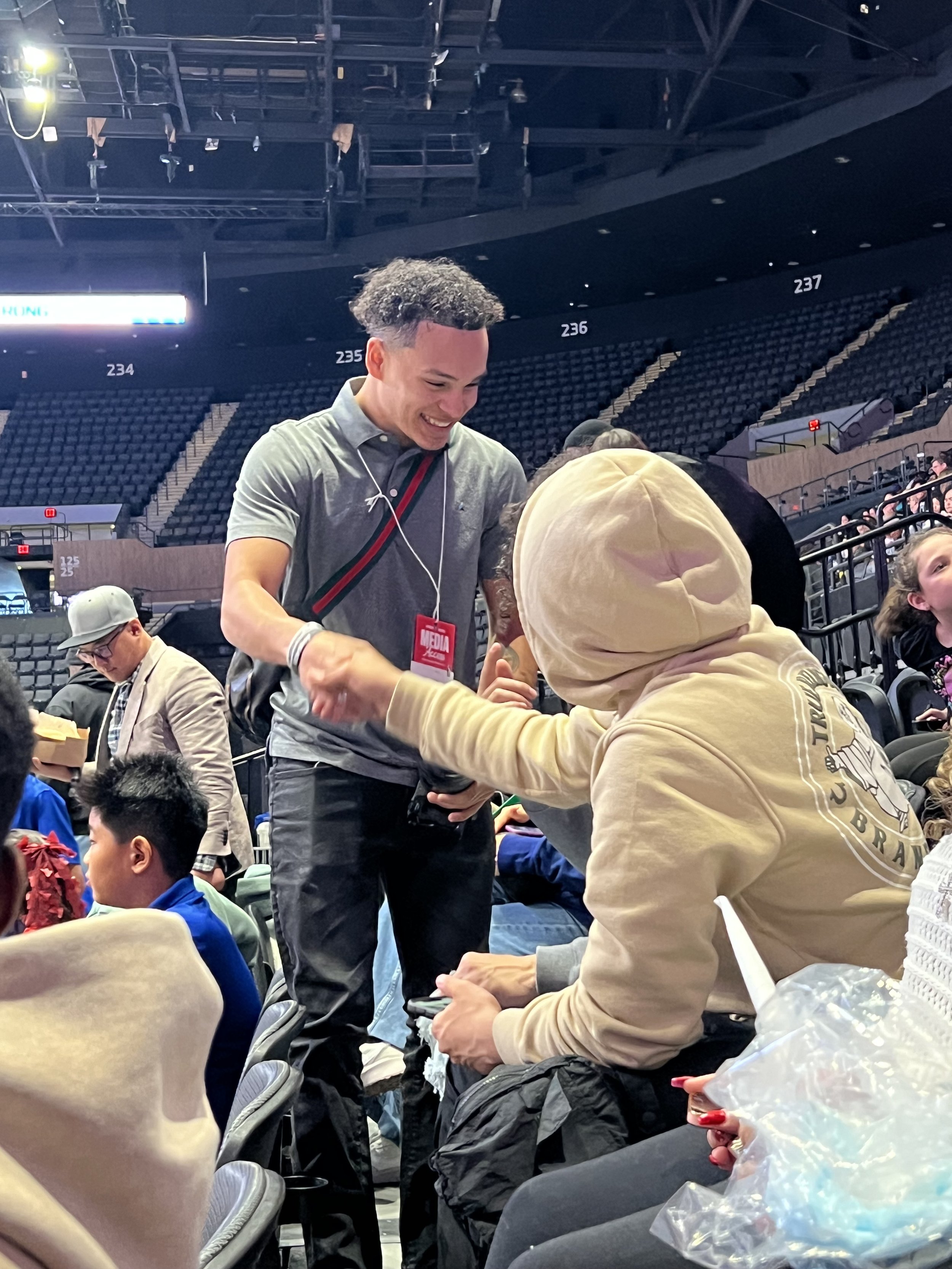 A young man with curly hair smiling and shaking hands with a person in a beige hoodie at an indoor event in an arena with empty seats. Several other people are seated, some wearing headphones, and there is a media badge on the young man.