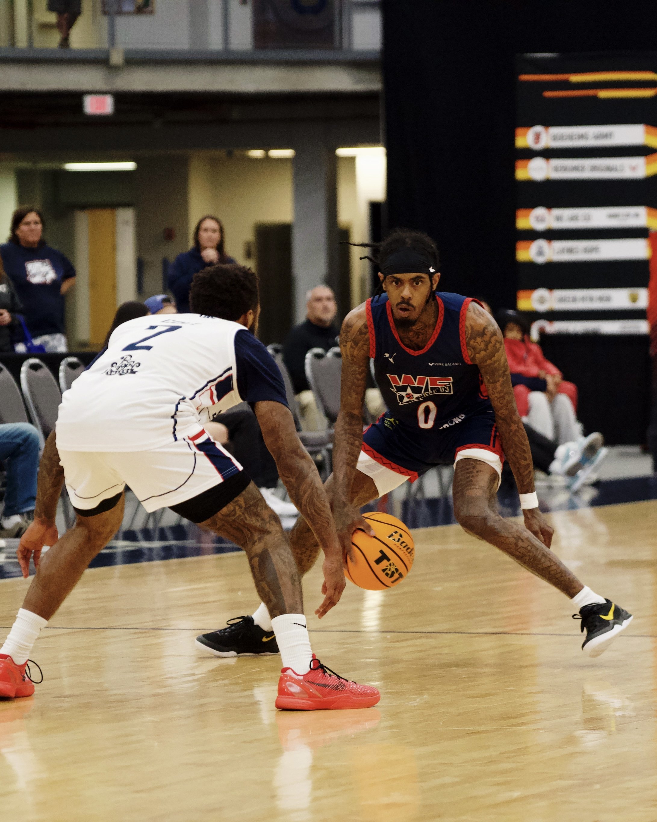 Two basketball players with tattoos and athletic attire are engaged in a game on an indoor court. One player in a black and red uniform is dribbling the ball, while the other in a white uniform is defending. Multiple spectators are in the background, some sitting and some standing.