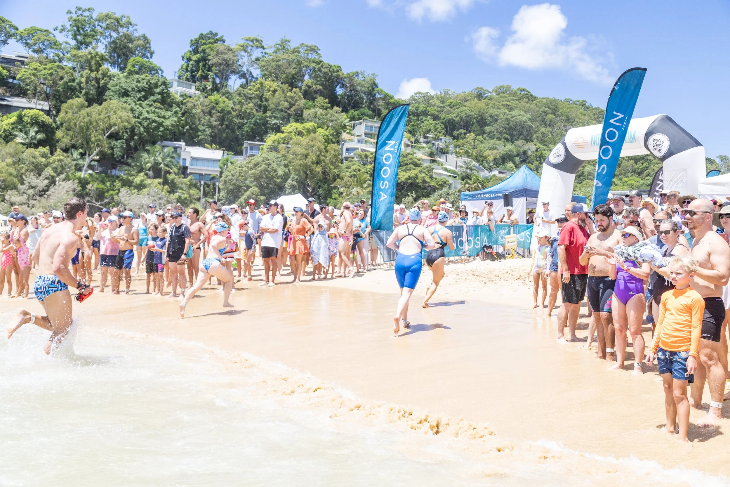 People participating in a swimming race on a beach, with spectators watching along the shoreline, banners, and a large inflatable arch in the background under a blue sky with clouds.