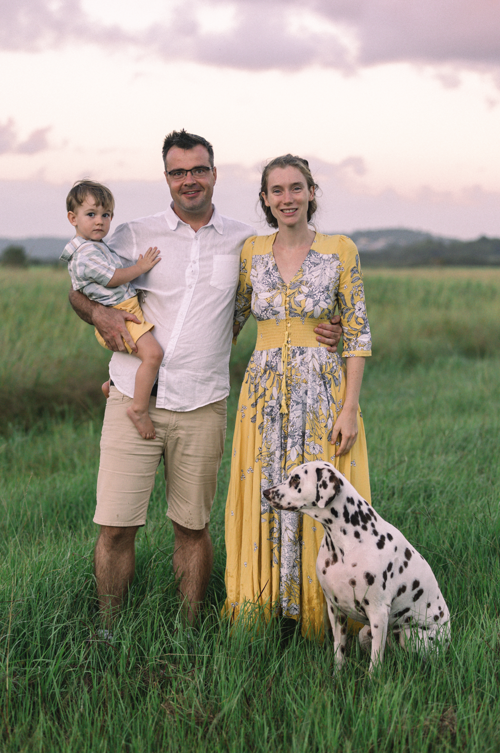A family of three standing in a grassy field with a Dalmatian dog, with a sky of pink and purple clouds at sunset.