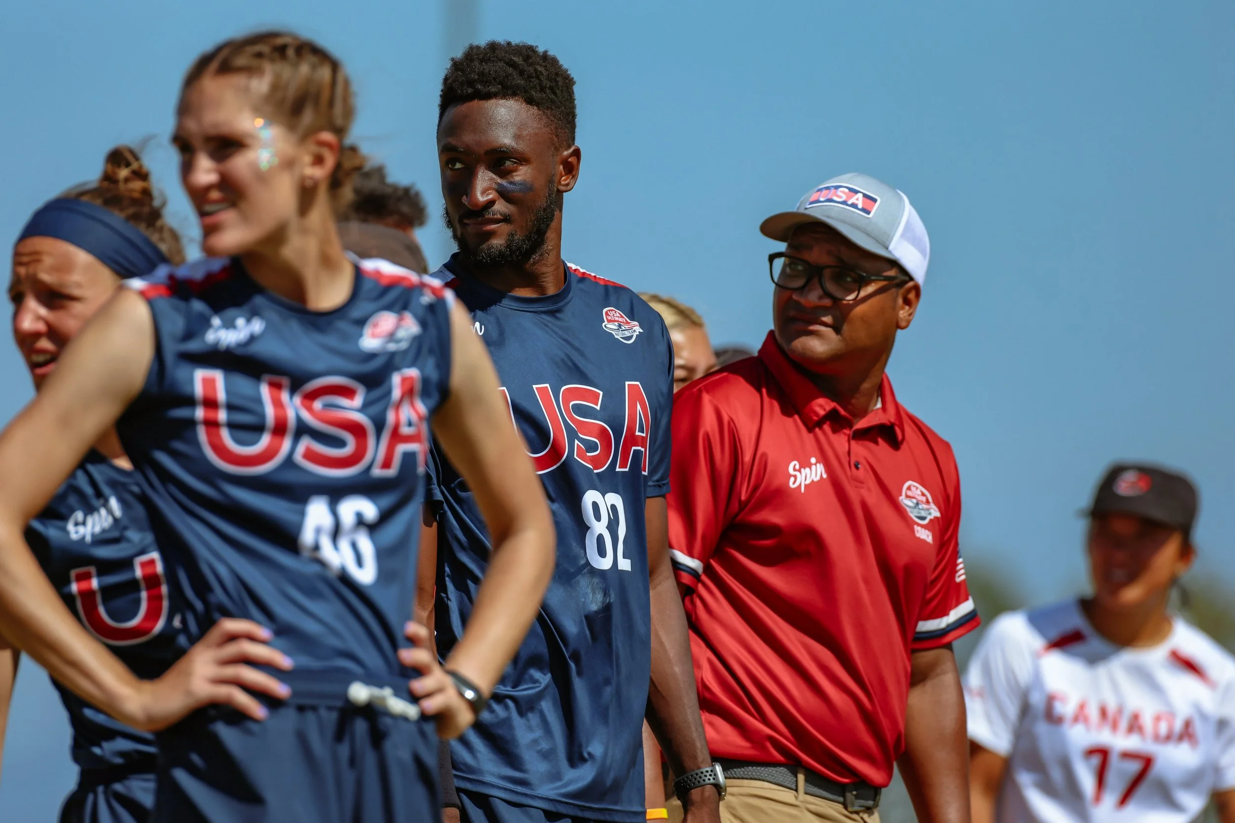 A group of athletes and a coach standing outdoors, wearing USA sports uniforms with the USA flag on their shirts, under a clear blue sky.