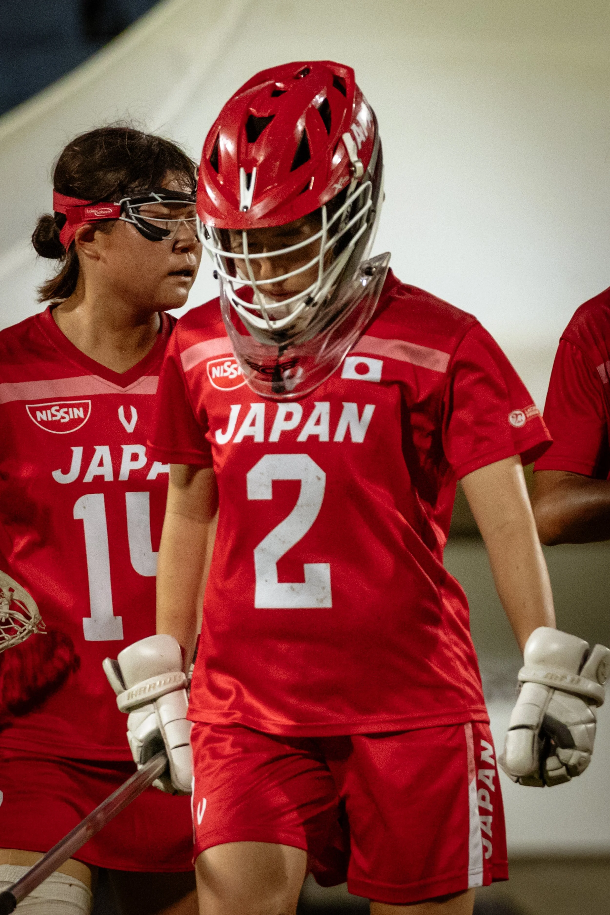 Japanese women's lacrosse players dressed in red jerseys with numbers 14 and 2, wearing protective gear and helmets, during a game or practice.