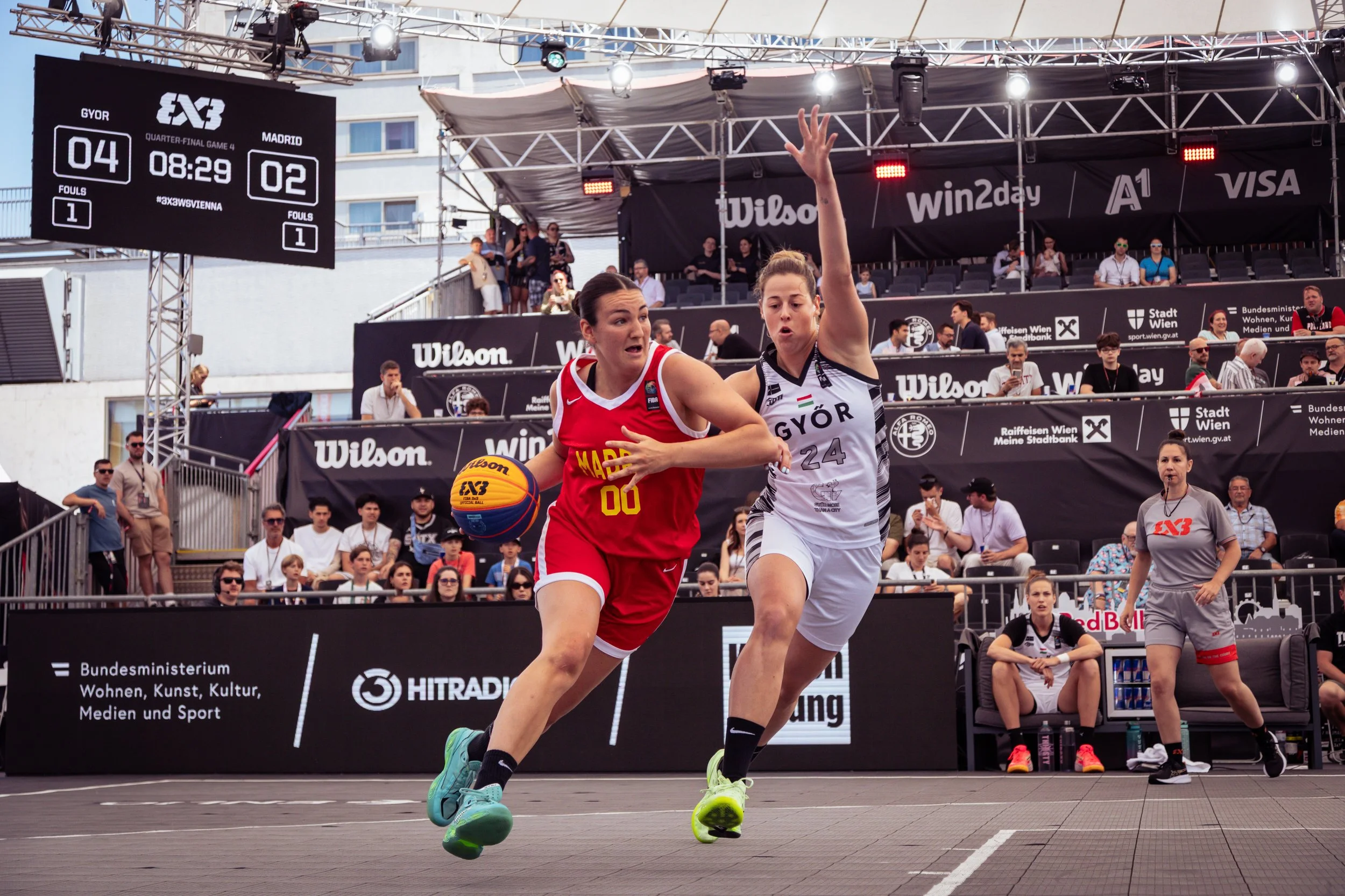 Two women playing basketball on an outdoor court during the 3x3 basketball Vienna World Tour, with spectators and officials watching, and a scoreboard showing the score and time.