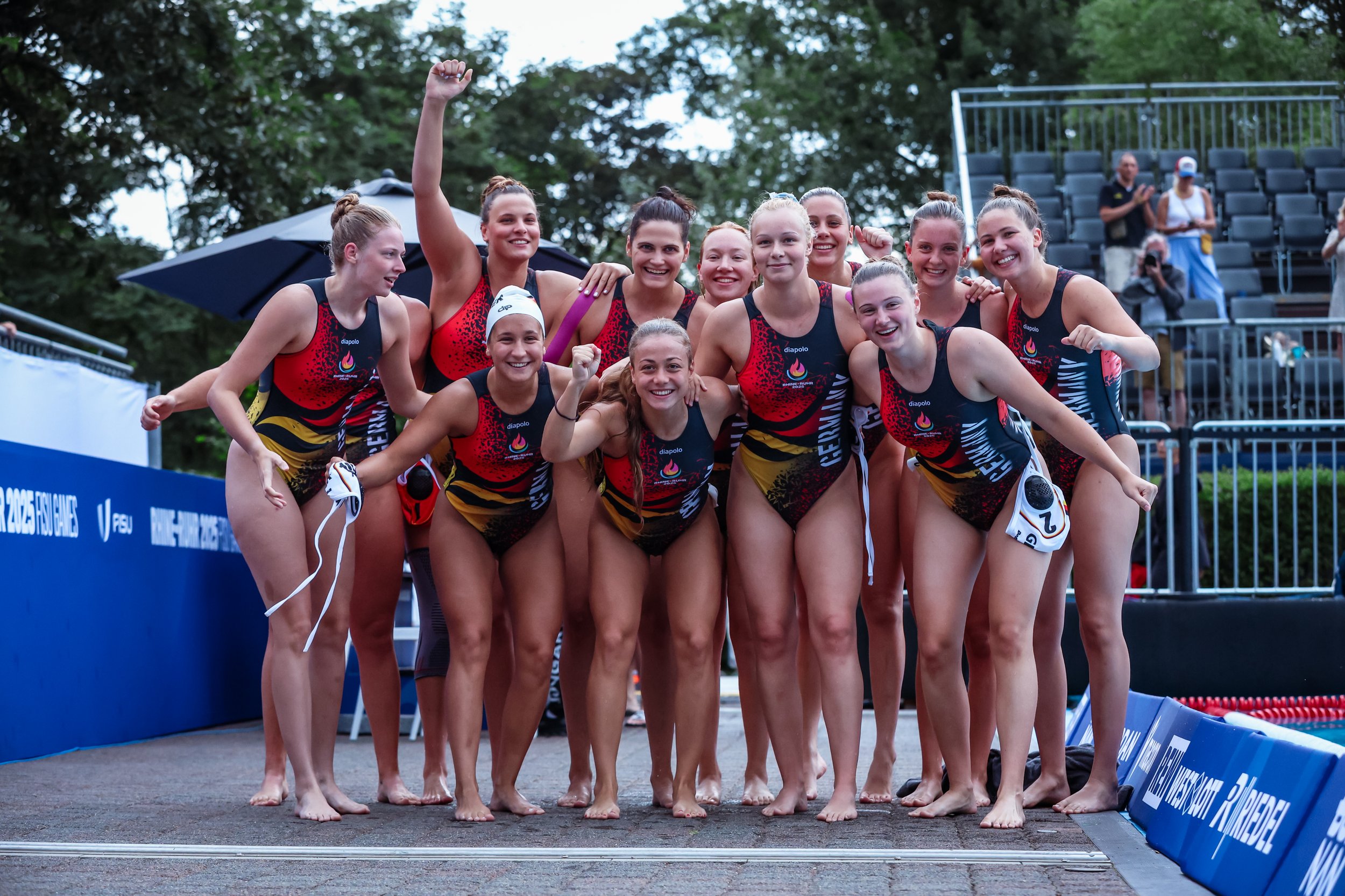 The German team celebrating together after a win at the World Uni Games 2025, wearing matching swimsuits with red, black, and yellow patterns, some raising fists or smiling.