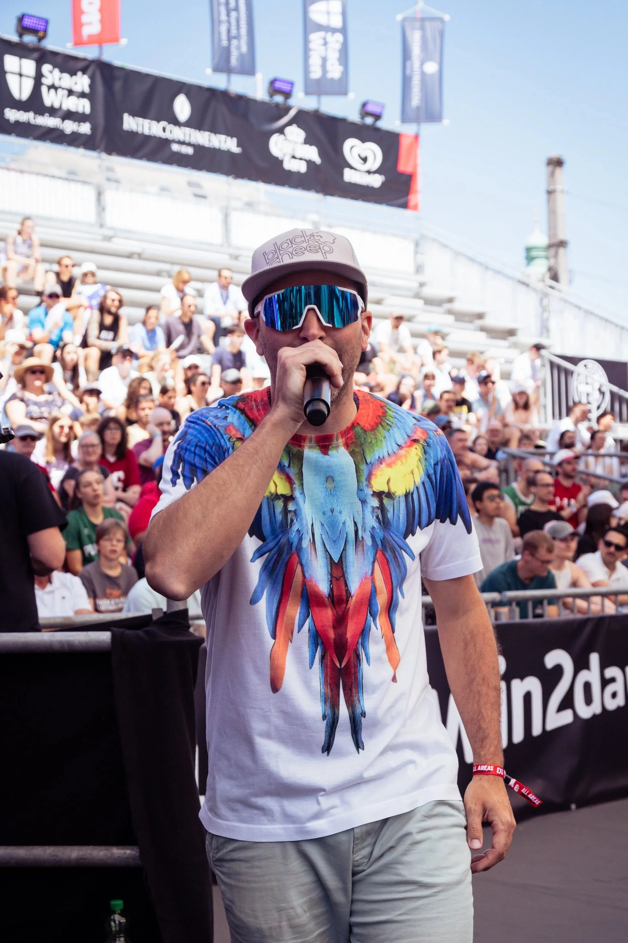 A man wearing reflective sunglasses, a beige cap, and a colorful parrot graphic T-shirt, holding a microphone at the 3x3 Vienna World Tour basketball event with a crowd of spectators in the background.