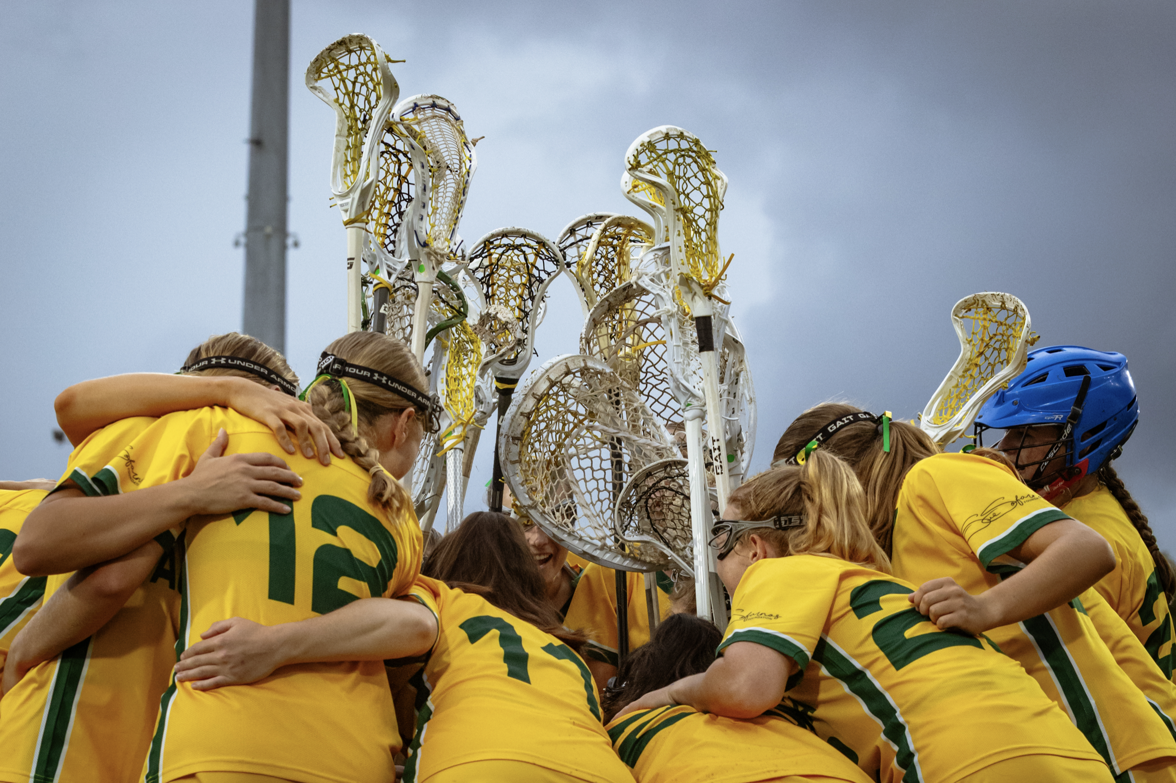 The women's Australian Lacrosse players in yellow uniforms huddled together in celebration, embracing each other, with their Lacrosse sticks raised in the air behind them, against a darkening cloudy sky.
