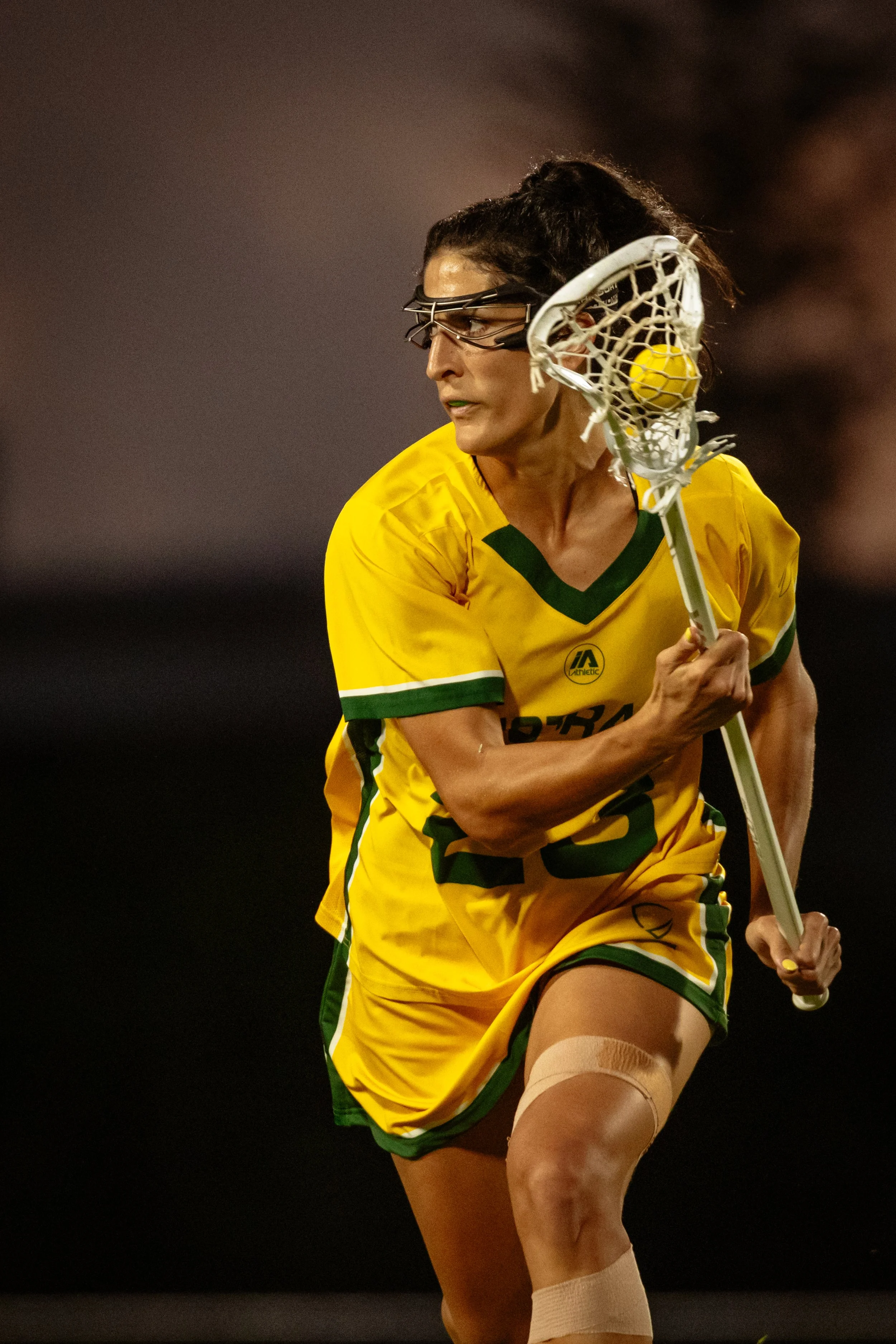 A female Australian lacrosse player in a yellow and green uniform with protective eyewear and a bandage on her thigh, holding a lacrosse stick with a ball in the net.