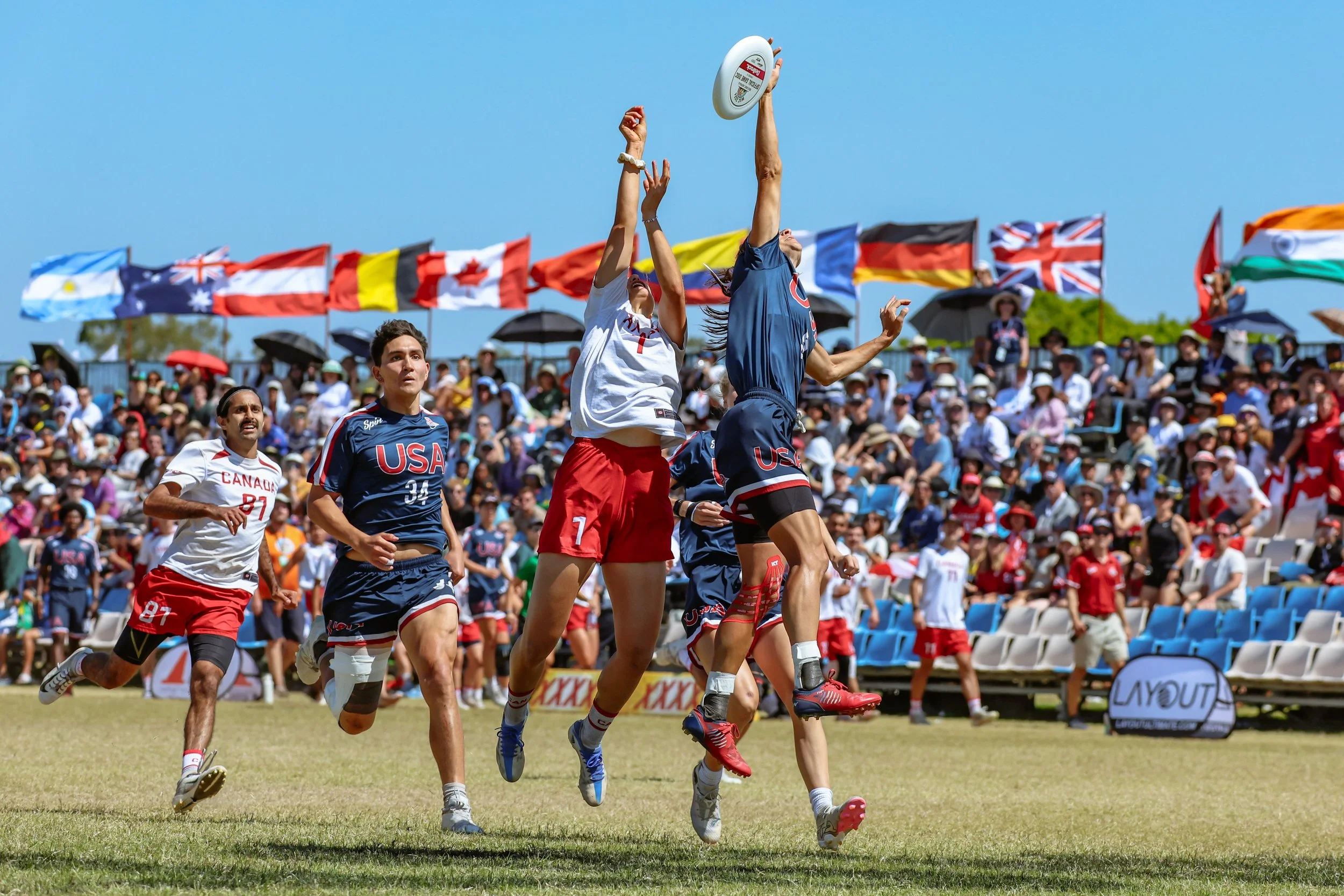 A mixed ultimate frisbee game in progress with players jumping to catch the frisbee on a grassy field, with spectators in the background and national flags flying. Taken at the World Ultimate Championships in 2024.