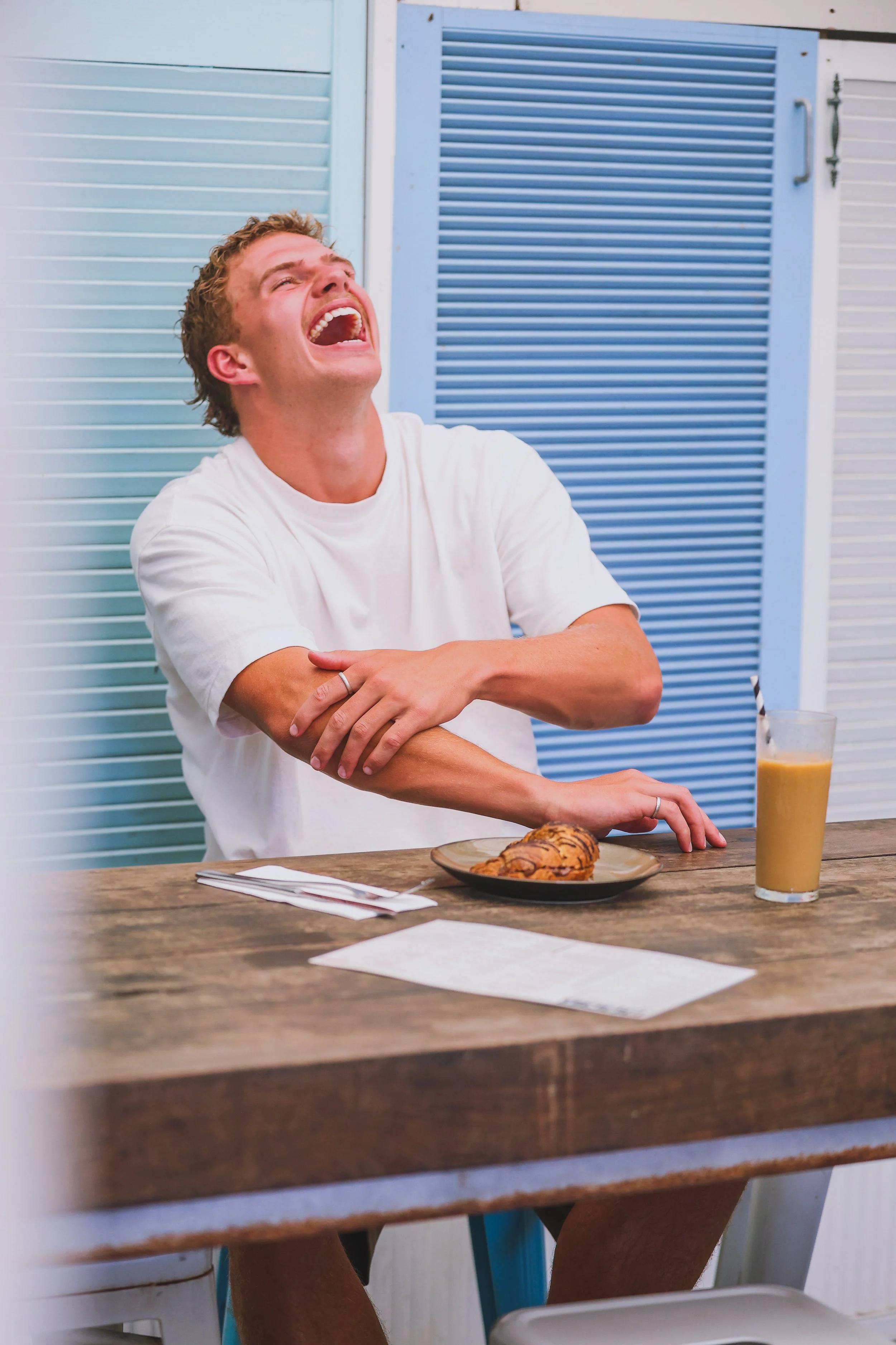 A young man sitting at a wooden table, laughing with his head tilted back and eyes closed, wearing a white t-shirt. There is a plate with a croissant and a glass of iced coffee on the table, with blue and white louvered shutters in the background.