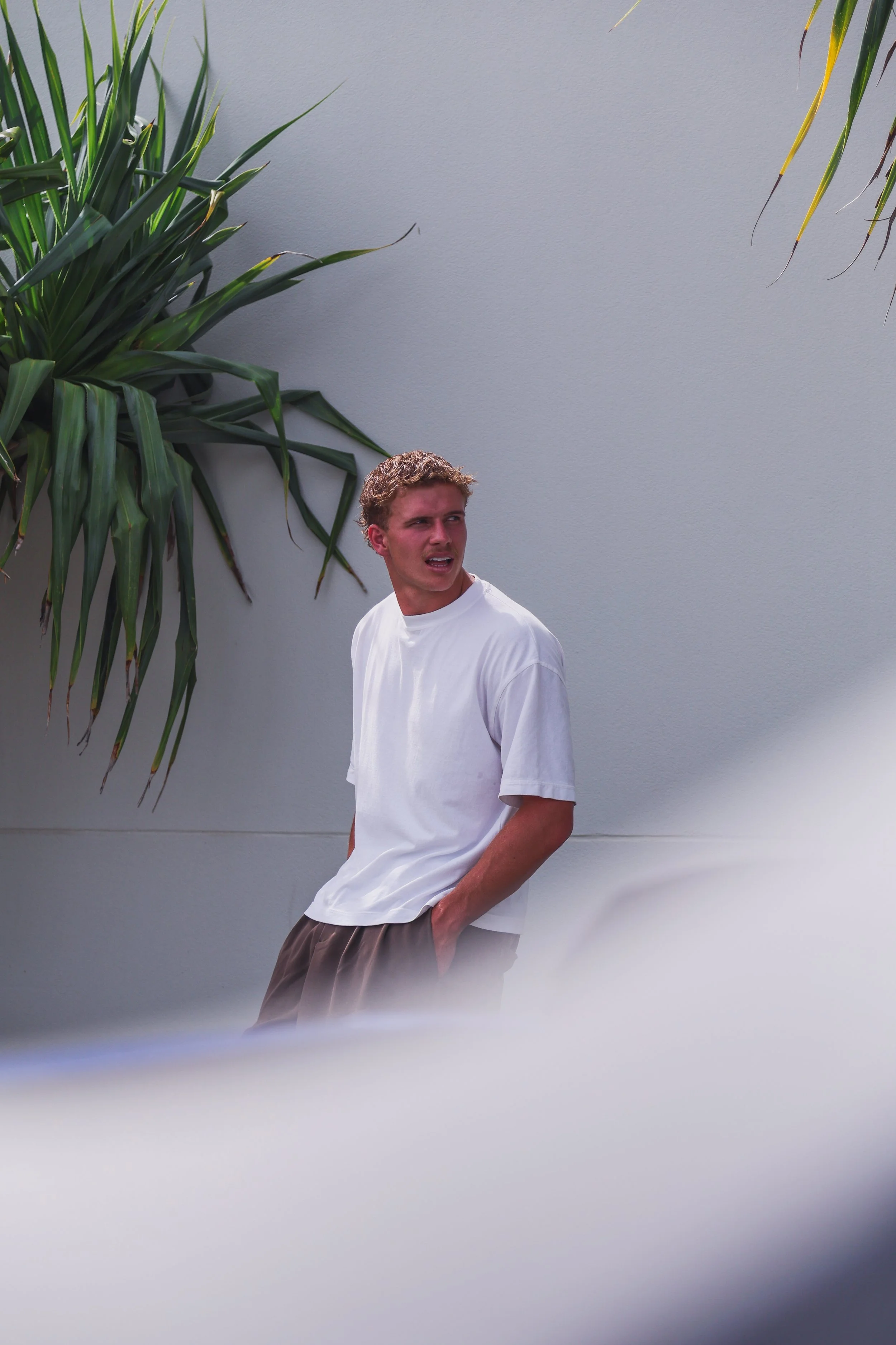 Gold Coast Sun's Jed Walker wearing a white t-shirt and brown shorts standing outdoors near a gray wall and large green plants, looking to the side with a concerned expression.