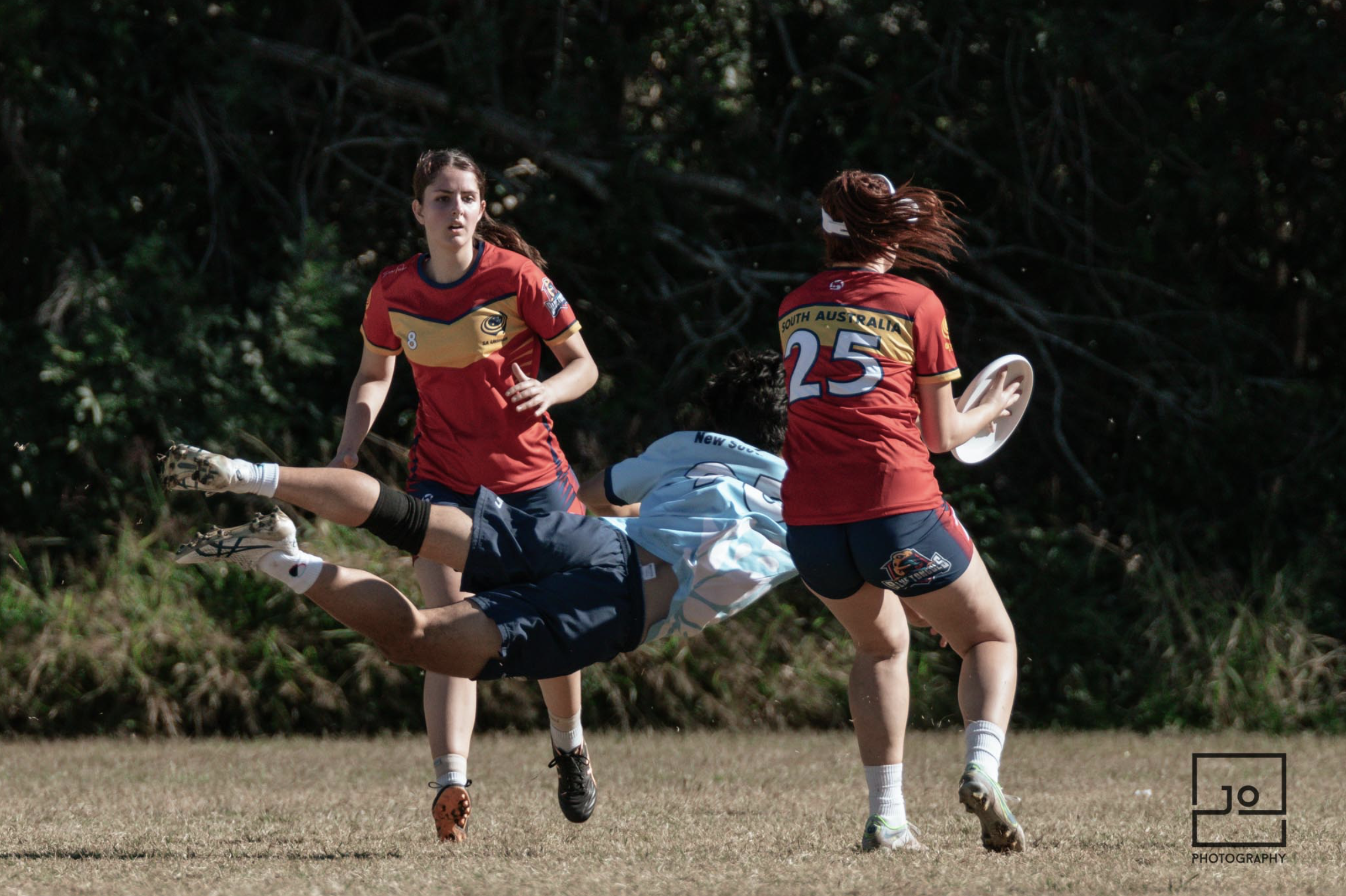 Two female rugby players in red and navy uniforms are on a grassy field, with one player in a white and light blue shirt diving forward while others look on, during a game.