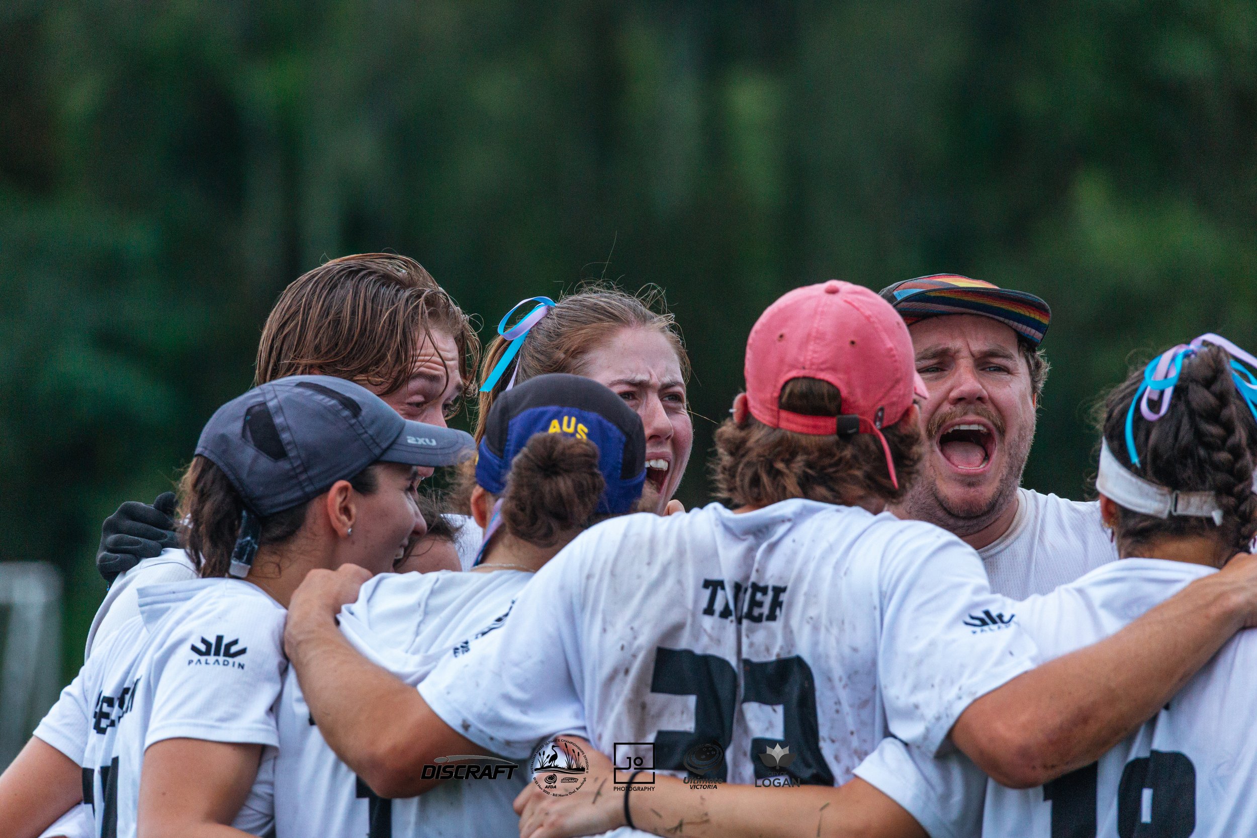 Group of athletes celebrating and hugging after a game of Ultimate Frisbee on a grassy field with trees in the background.