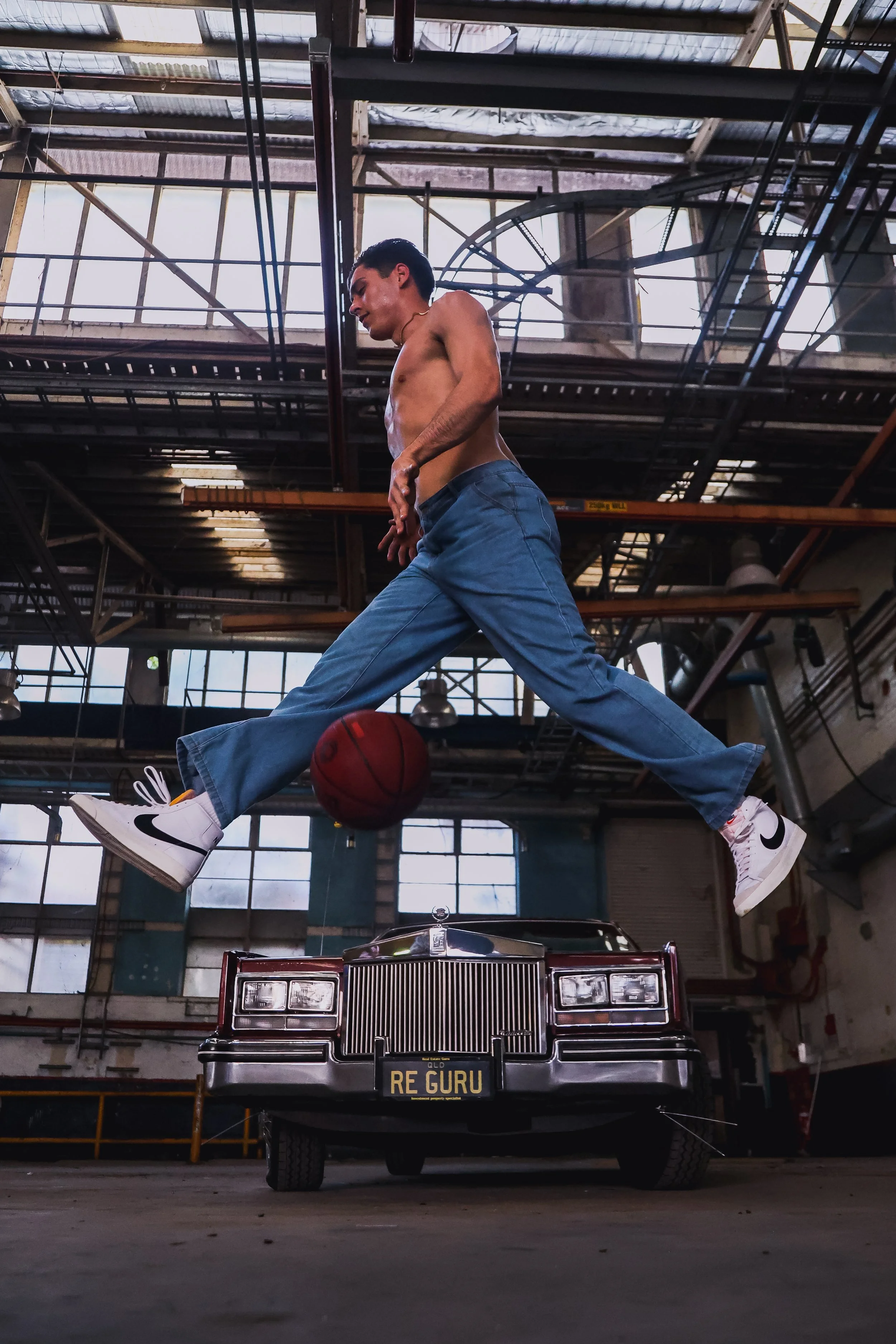 A shirtless man jumping in the air with a basketball, wearing white Nike sneakers, in an industrial garage or warehouse, with a vintage black car with a customized license plate that reads 'RE GURU' underneath him.