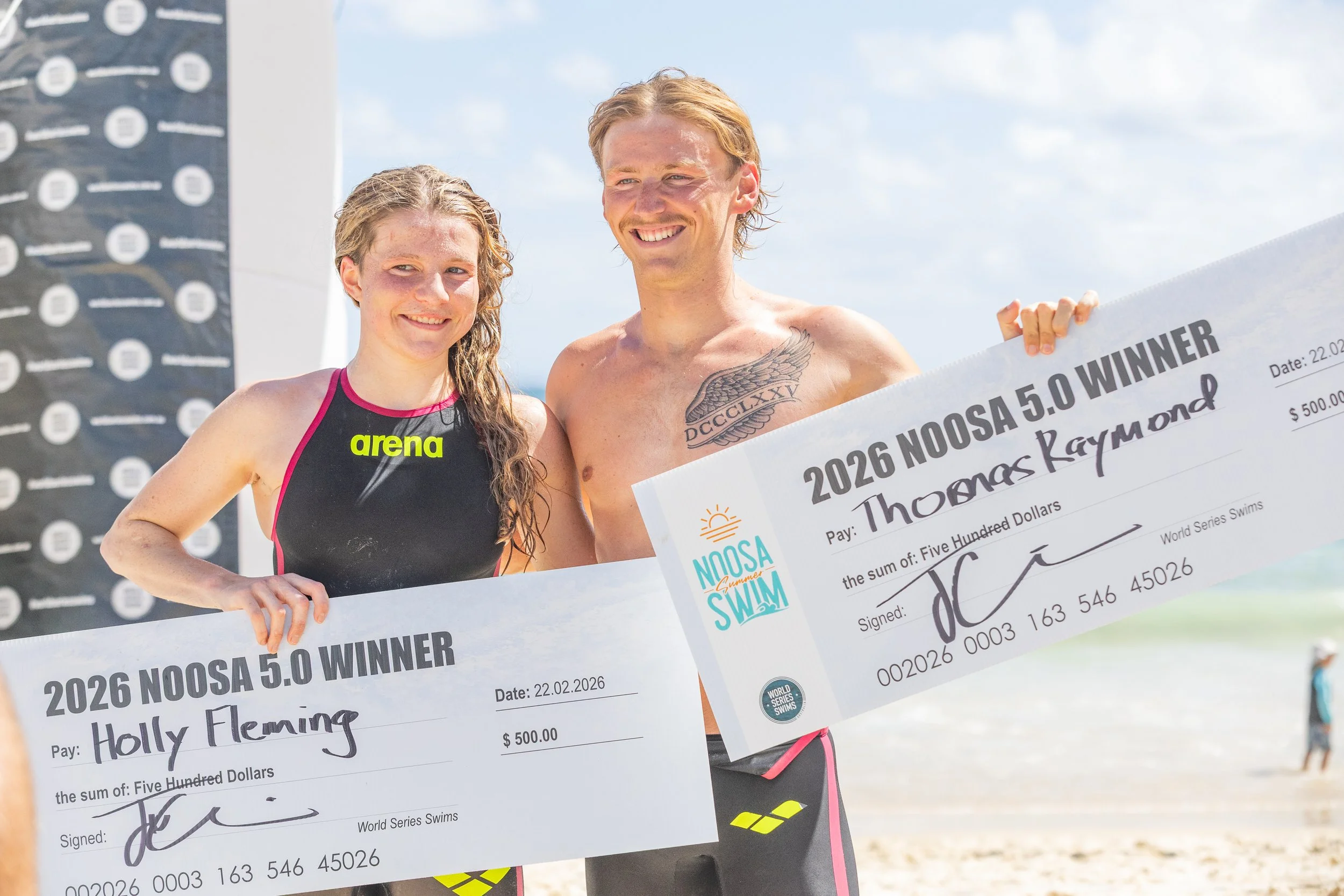 Two young swimmers, a woman and a man, holding large checks for winning a swim competition, smiling at the camera on a beach.