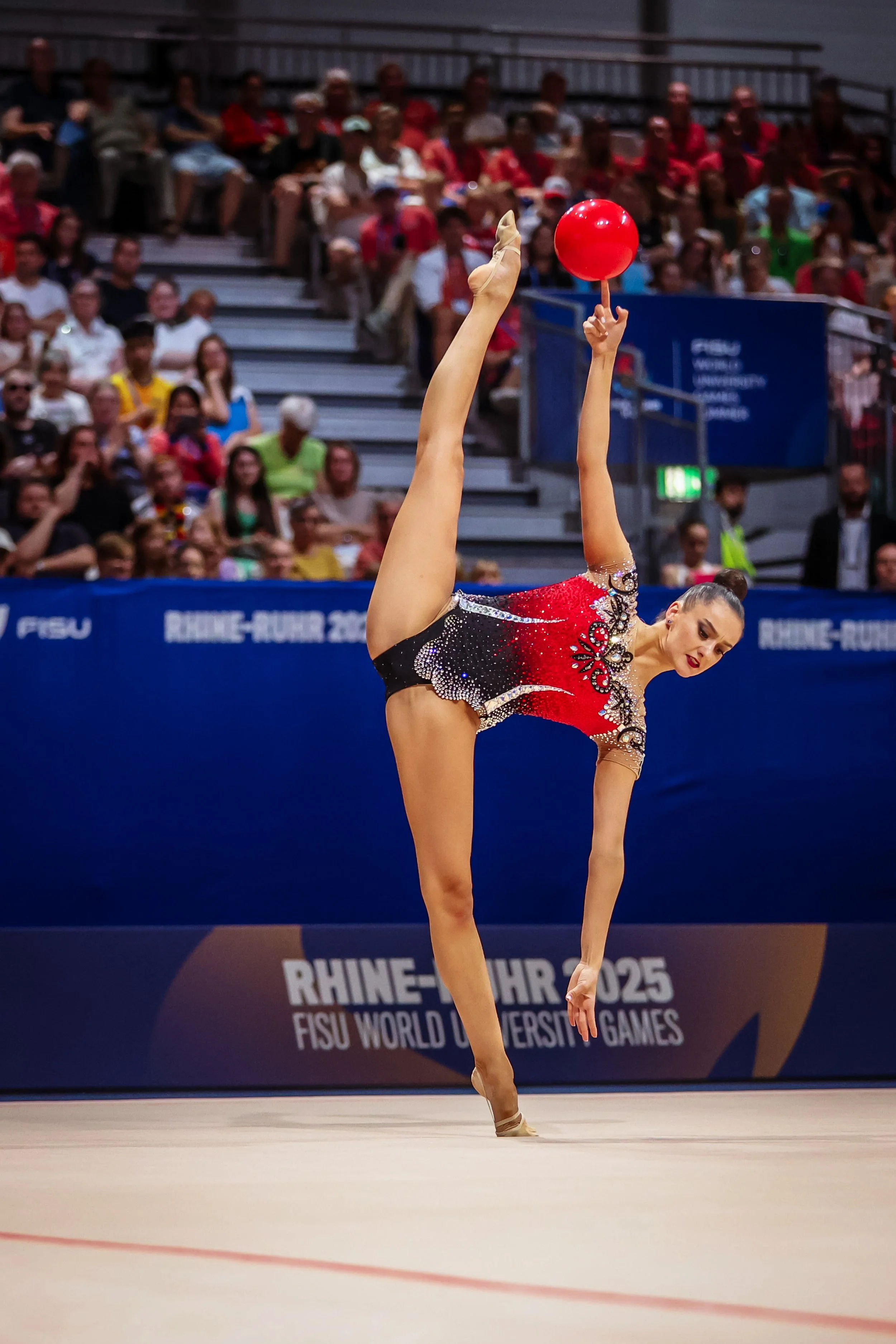 A rhythmic gymnast in a red and black leotard performing a stunt with a red ball at the Rhine-Ruhr 2025 FISU World University Games, with an audience watching from the bleachers.