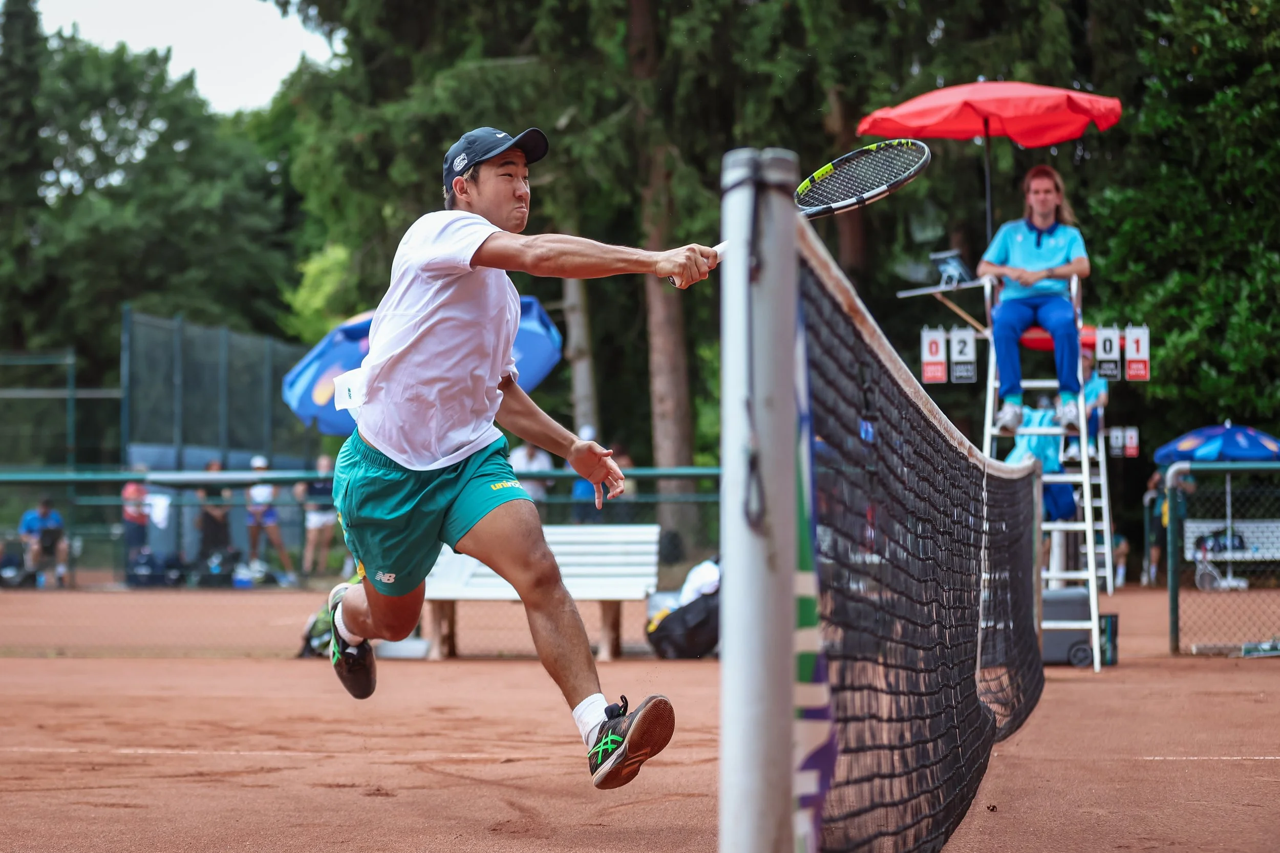 Australian tennis player in a white shirt and teal shorts hitting a tennis ball near the net on a clay court during a match, with a female umpire sitting on a chair under a red umbrella in the background. Taken during World Uni Games 2025 in Germany.