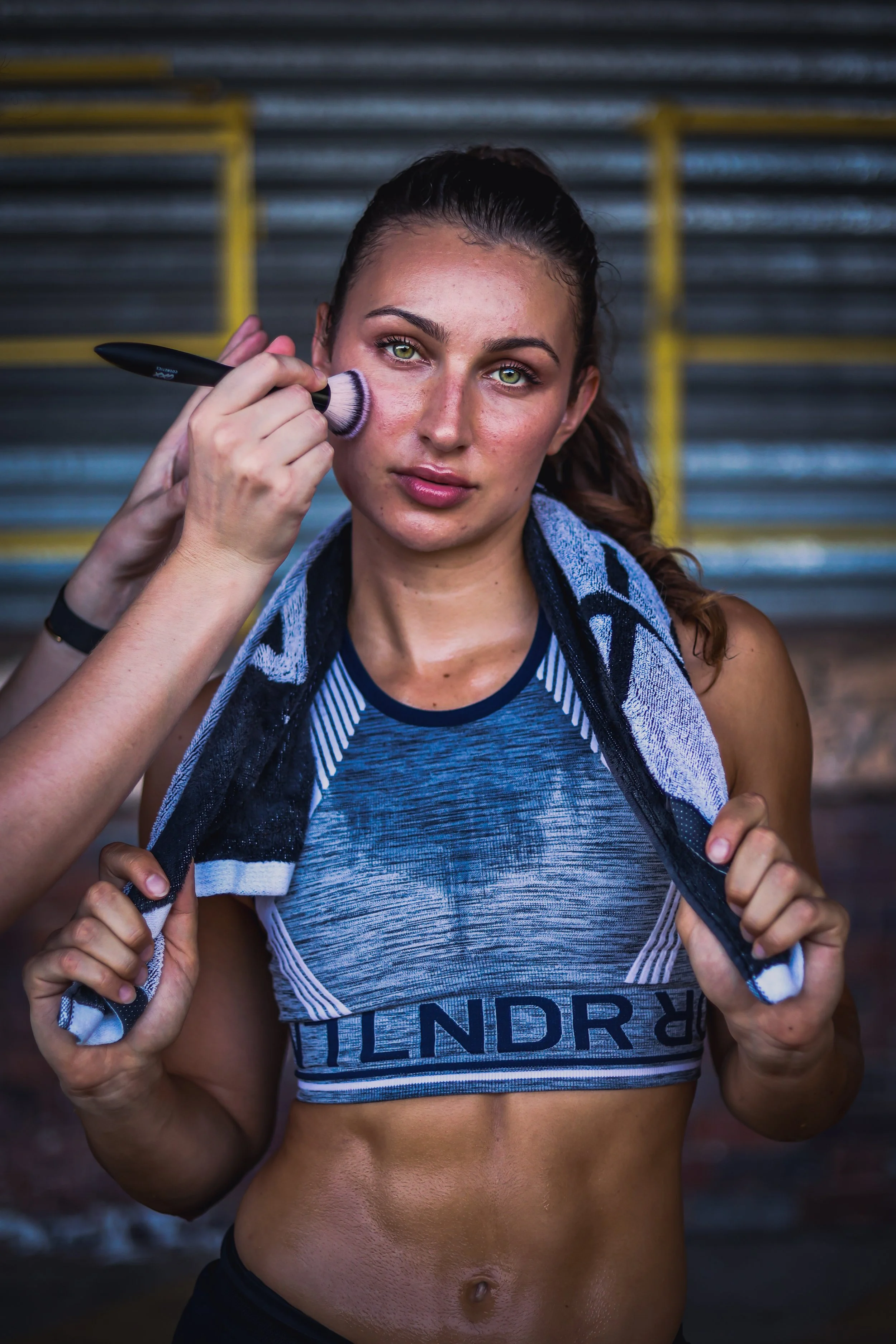 Zoe Isabella with sweat on her face and athletic clothing is having makeup applied to her cheek with a makeup brush, holding a towel around her neck, standing in front of metal bleachers. Athlete photoshoot.