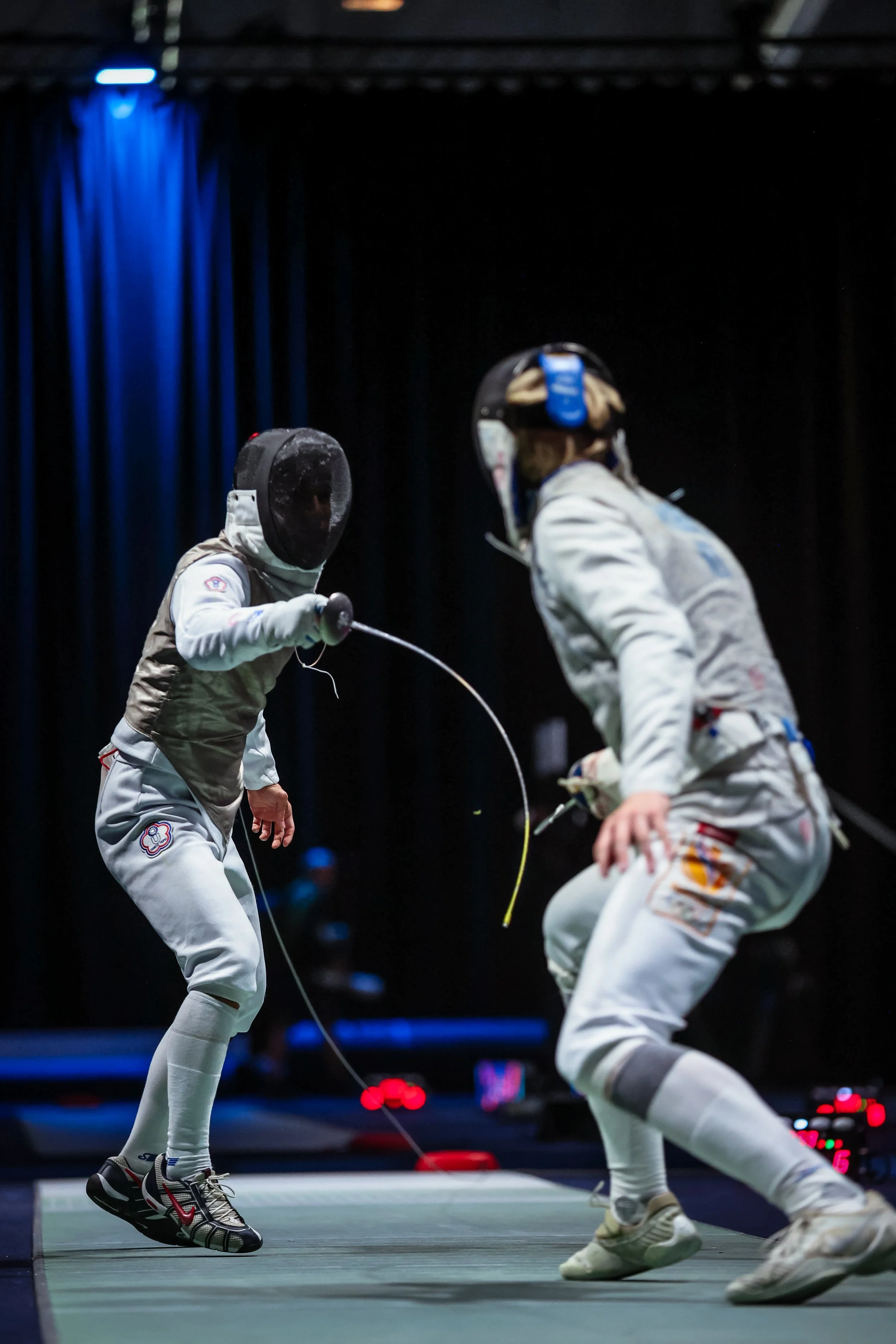 Two fencers in protective gear fencing on an indoor piste, with a dark background and blue stage lighting.