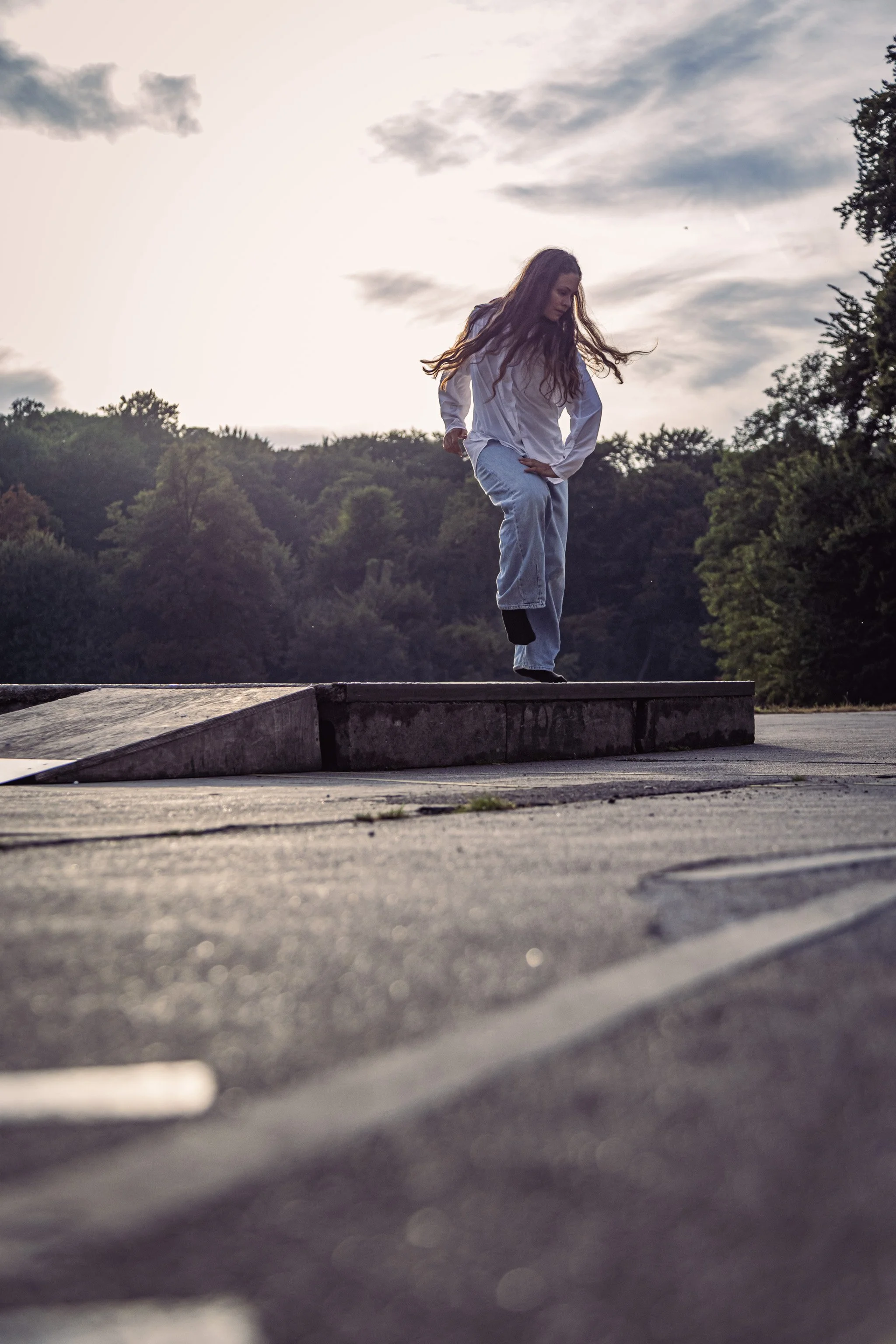 A young woman with long hair, dressed in a white outfit, balances on a curb in an outdoor setting at sunset, with trees and a cloudy sky in the background.