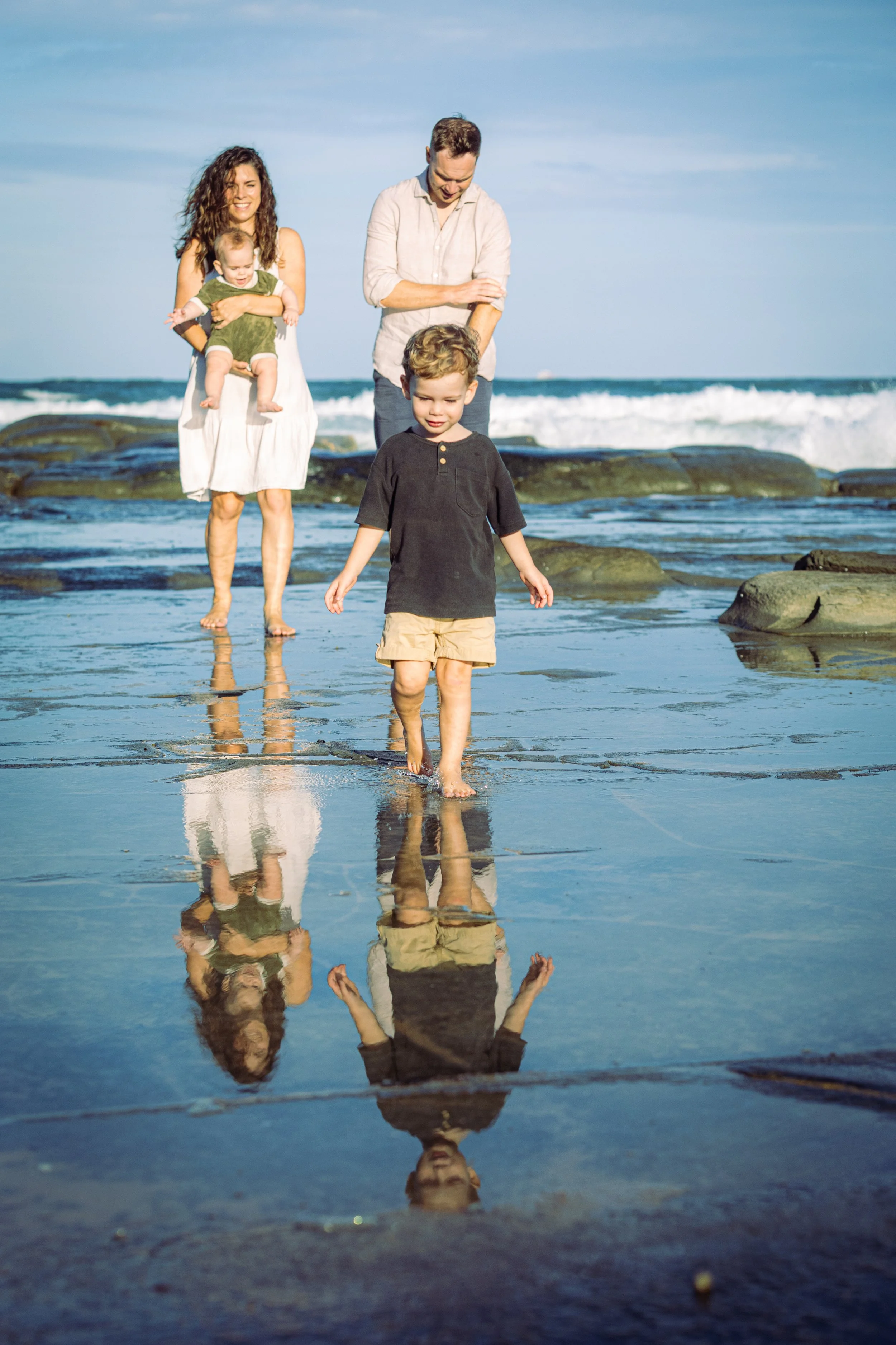 A family walking on the beach during the daytime, with the ocean in the background. The mother is holding a young child, and an older boy is walking in front, all barefoot on the wet sand that reflects them.