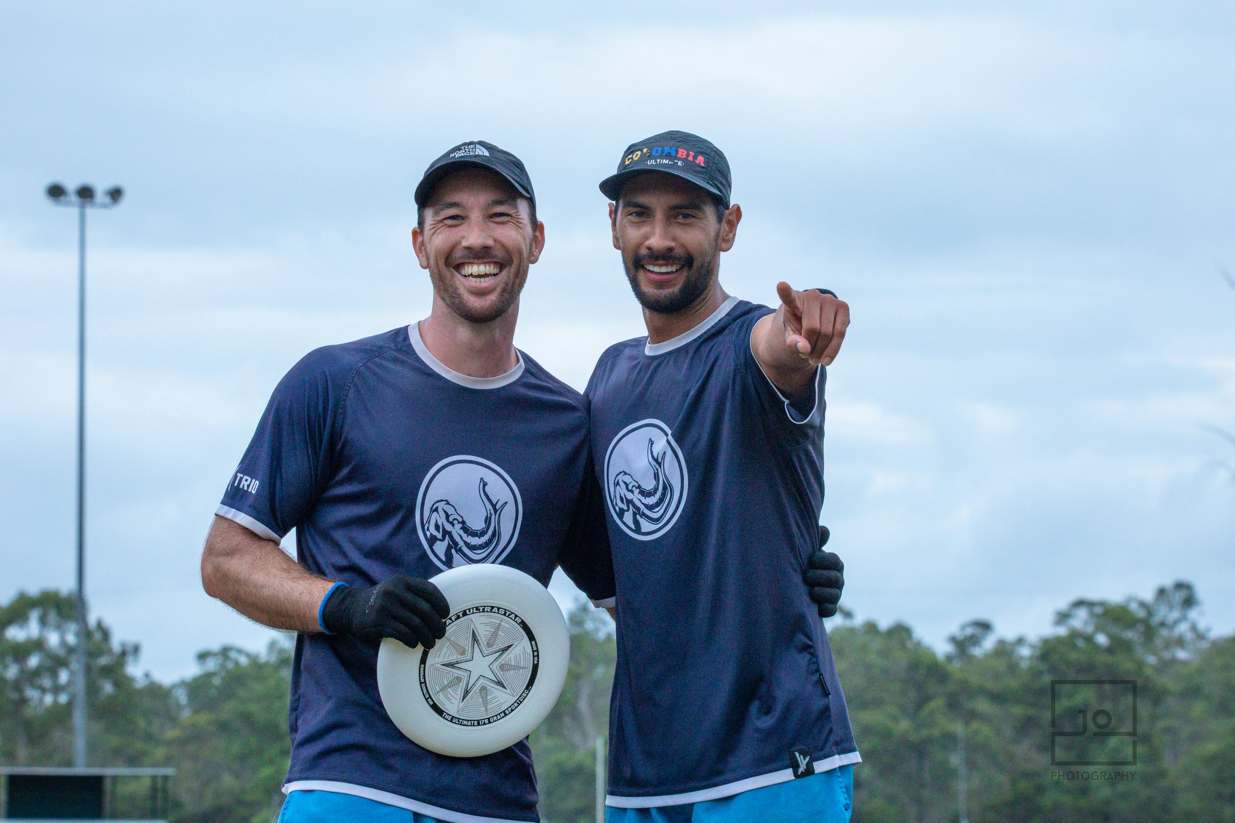 Two smiling men in matching blue sports jerseys and caps, one holding a disc and the other pointing at the camera, standing on a field with trees and cloudy sky in the background.