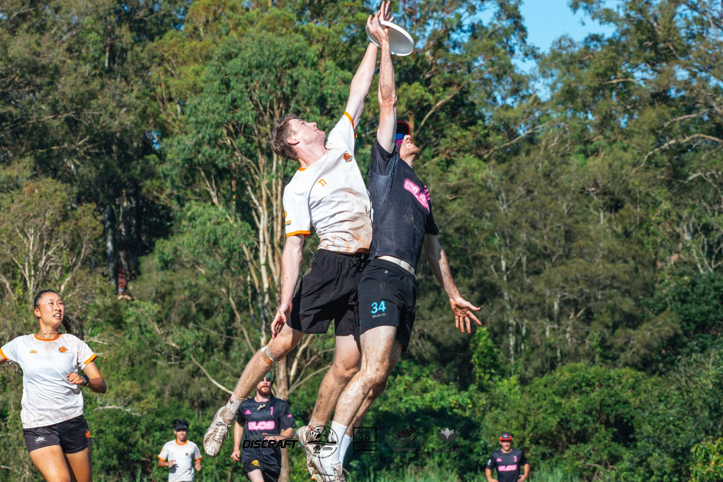 Two men are jumping to catch a frisbee during an Ultimate Frisbee game, with spectators and forest in the background.