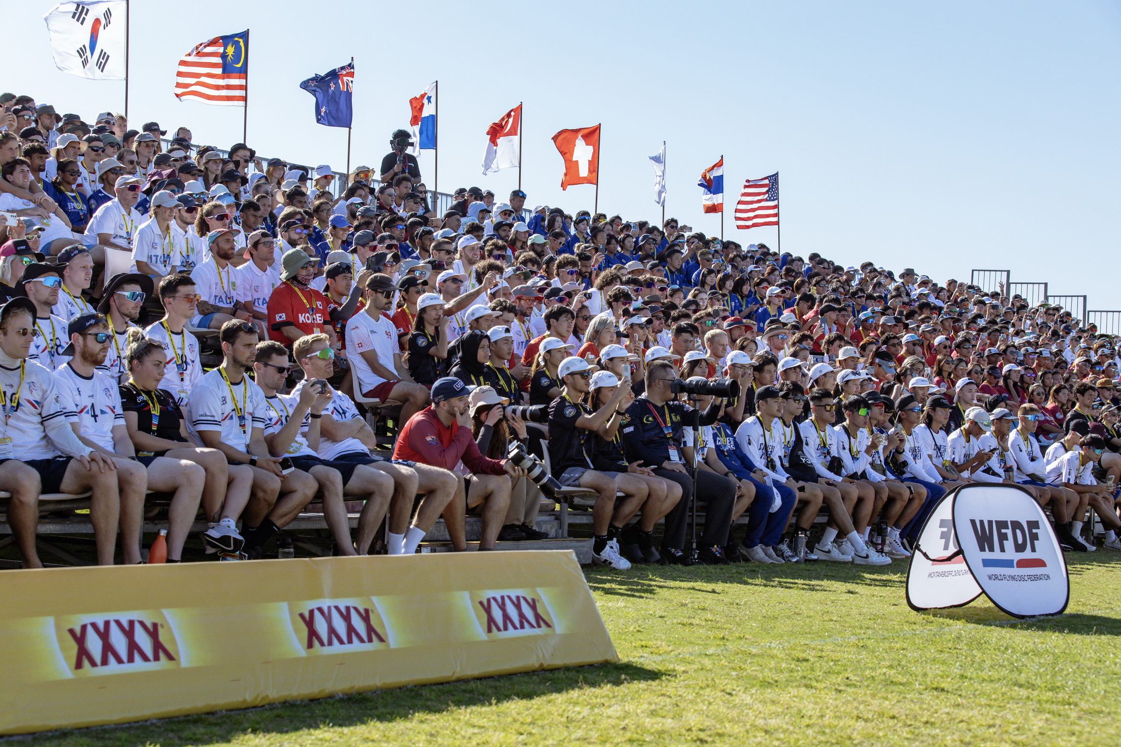 Crowd of spectators watching the World Ultimate Championship event, seated on bleachers under a clear blue sky, with numerous international flags flying overhead and banners on the ground displaying logos.