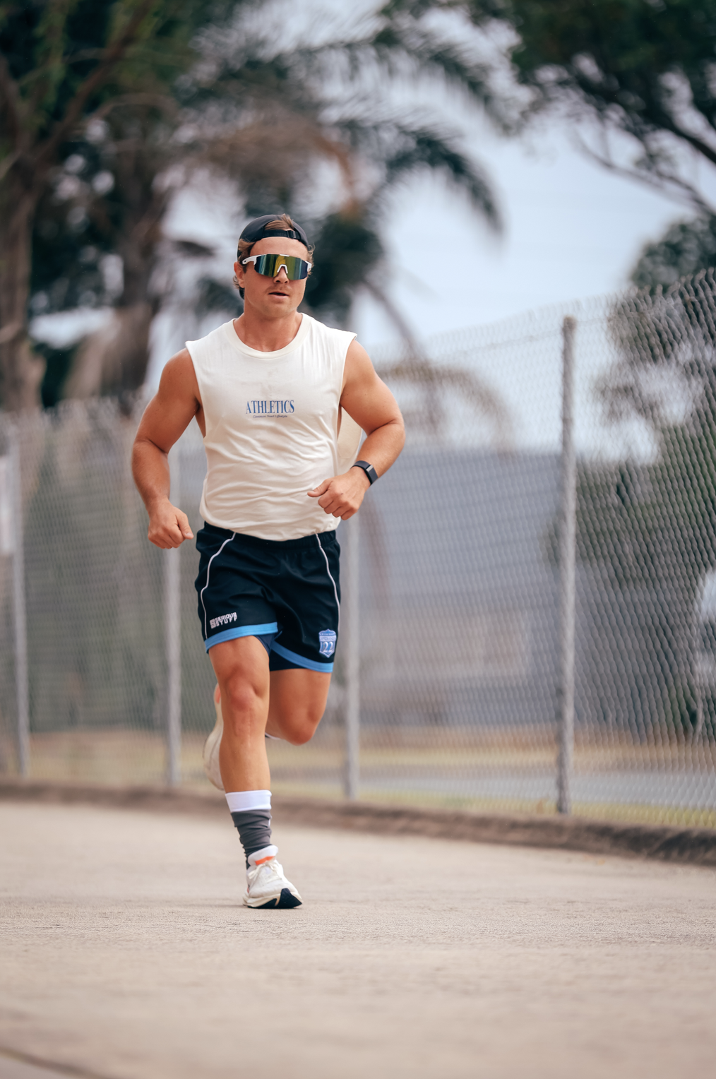 Nick Luff running on a paved outdoor path, wearing athletic shorts, a sleeveless shirt, sunglasses, and a watch, with a chain-link fence and trees in the background.
