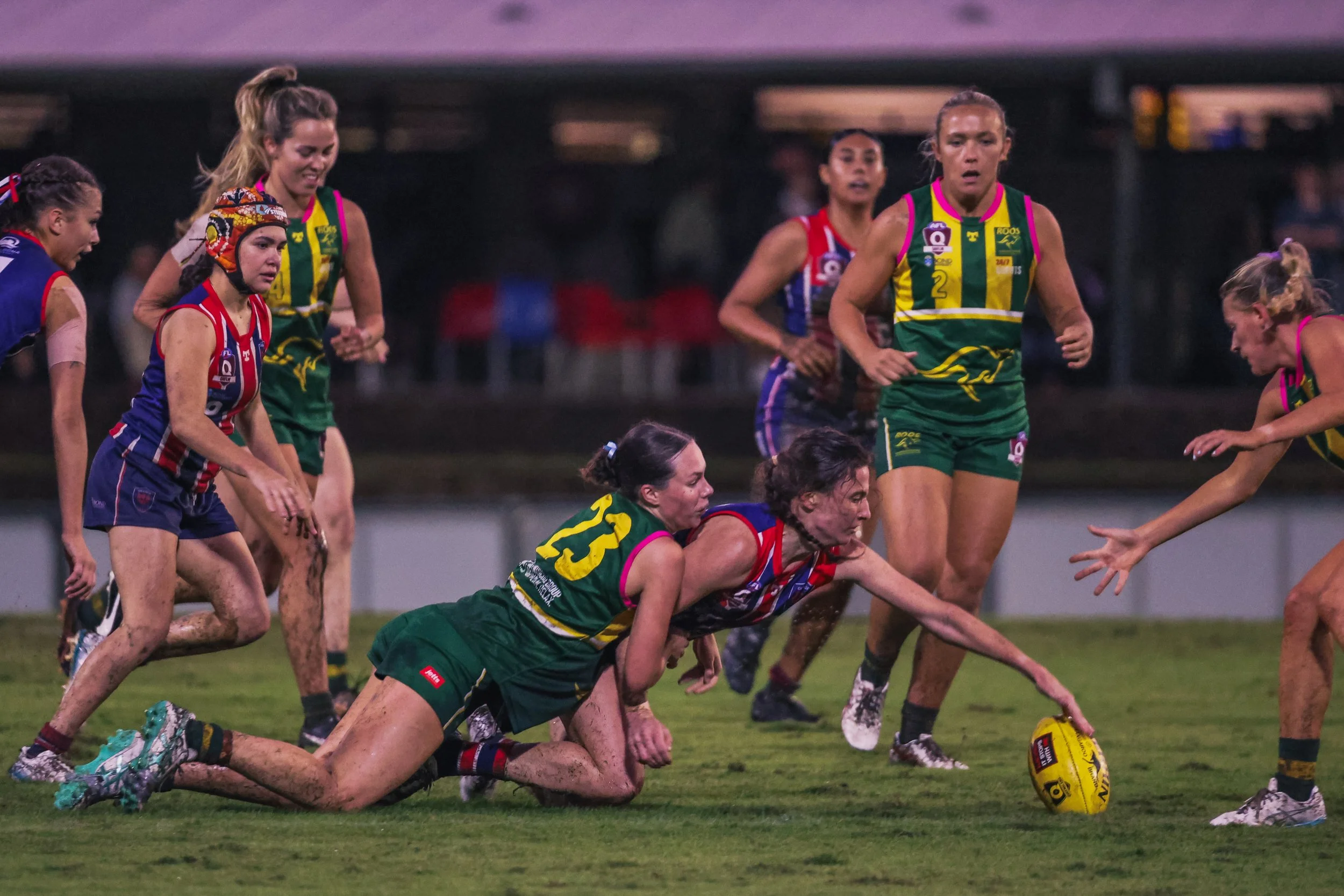 Women playing Australian rules football on a muddy field during a game, with some players reaching for the ball and others falling to the ground.