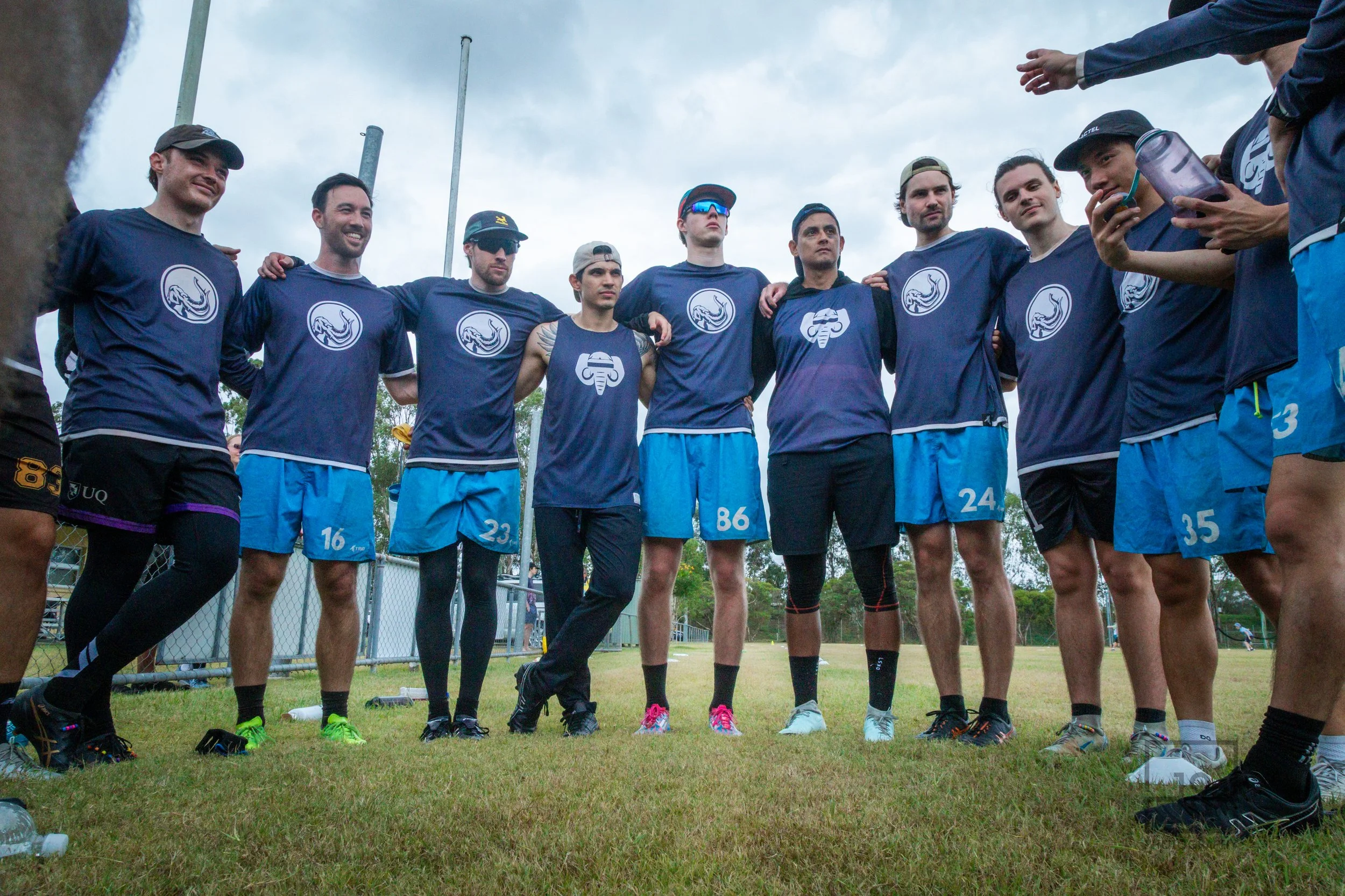 A group of Ultimate Frisbee Players standing in a semi-circle on a grass field, wearing matching blue sports jerseys and shorts, the Mammoth Ultimate Club uniform, with their arms around each other's shoulders, team huddle during the QLD Regionals