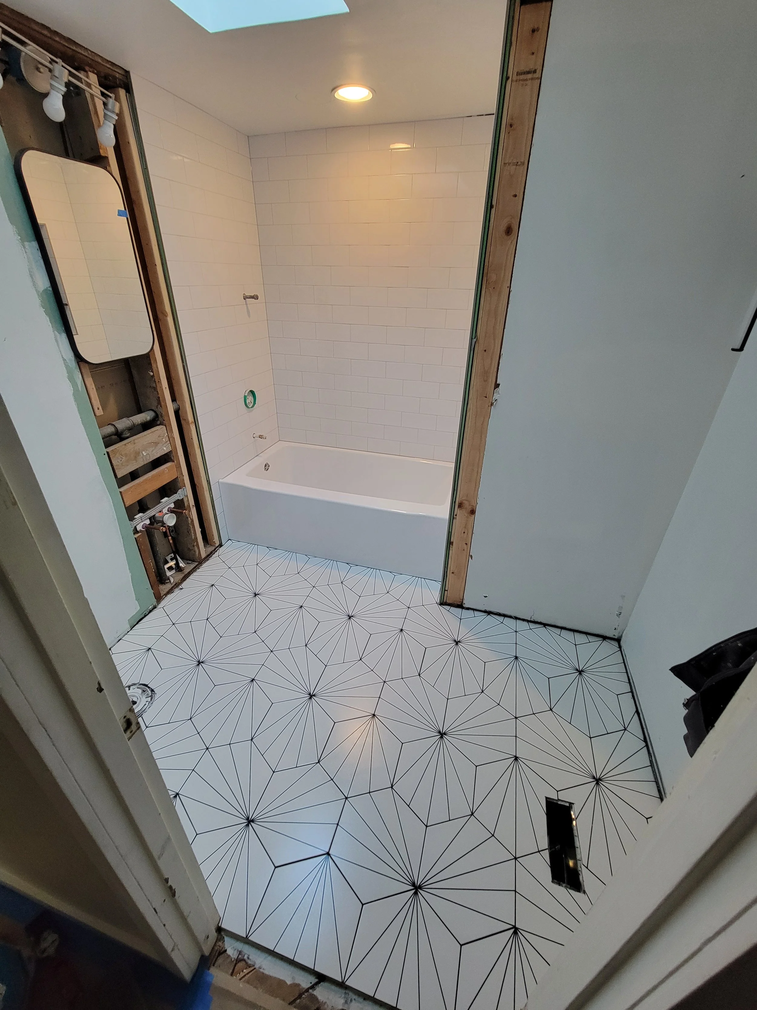Bathroom under renovation with geometric patterned floor tiles, white tiled walls around a bathtub, and exposed wood framing.