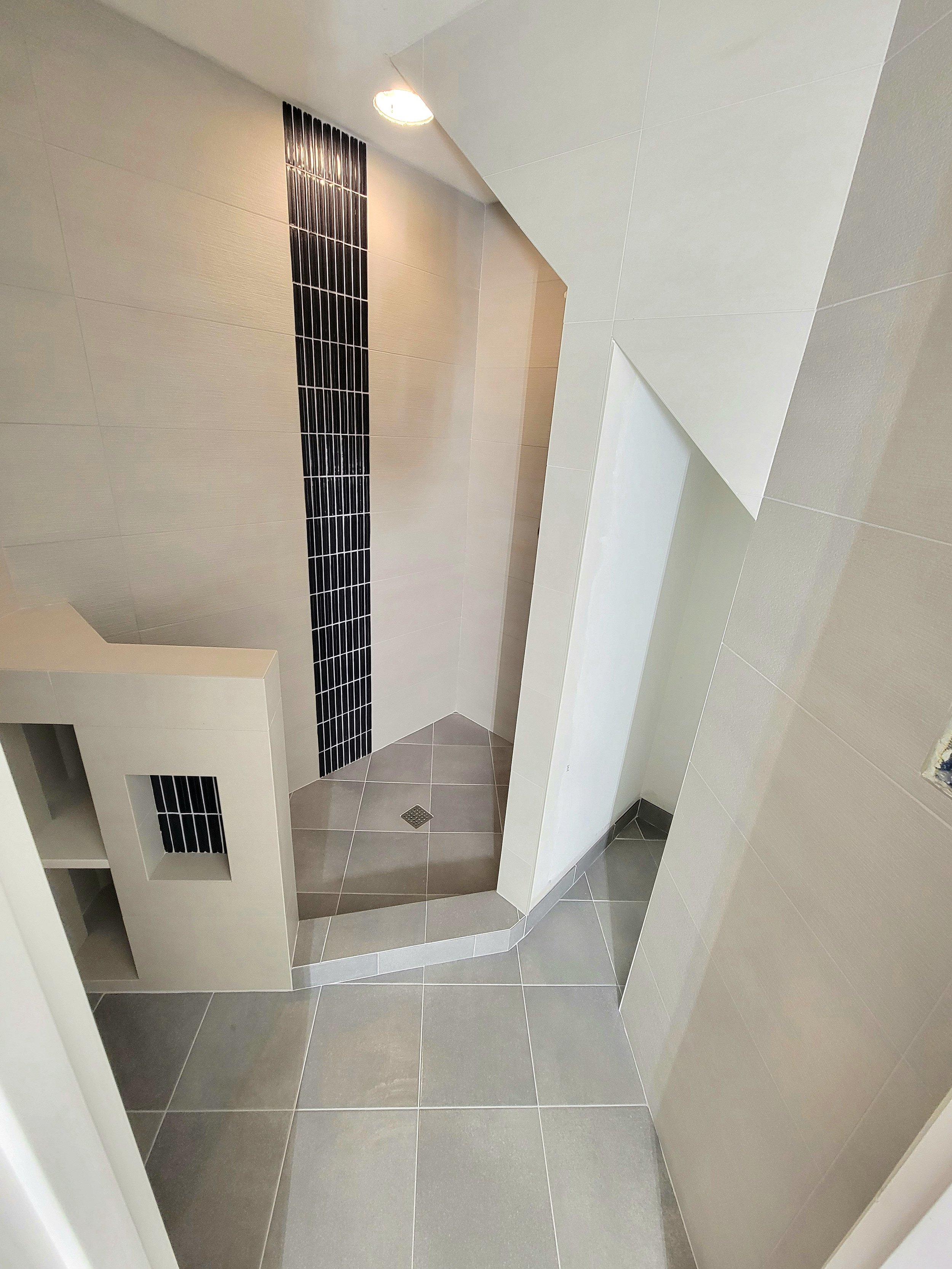 A modern shower area with beige and black tiled walls, a small raised tiled section with a drain, and a partial wall dividing it from the rest of the bathroom.