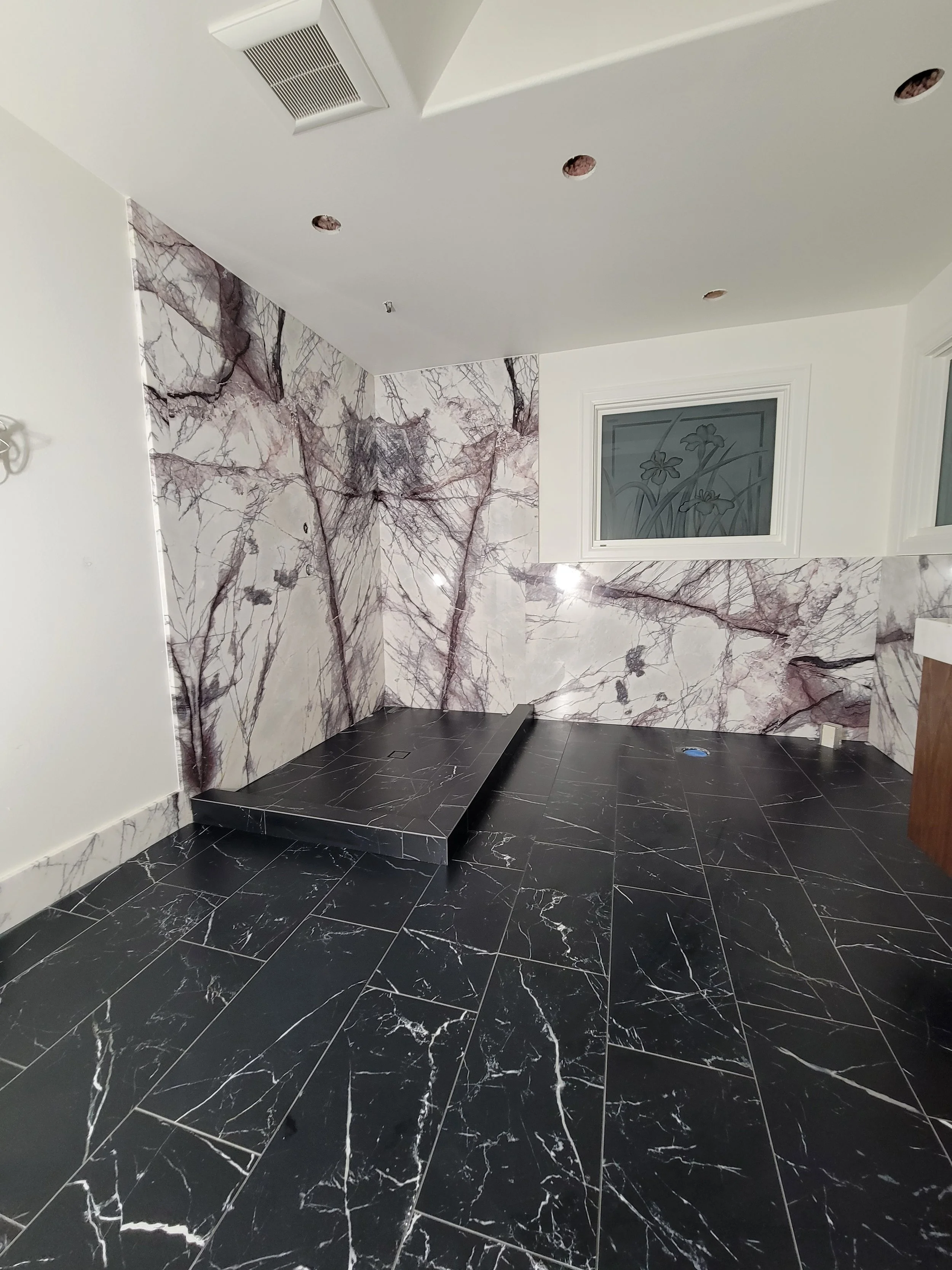 Room with black marble floor and marble walls featuring pink and gray veining, and a small raised platform. There are two windows with decorative floral frosted glass. Ceiling has recessed lights and an air vent.