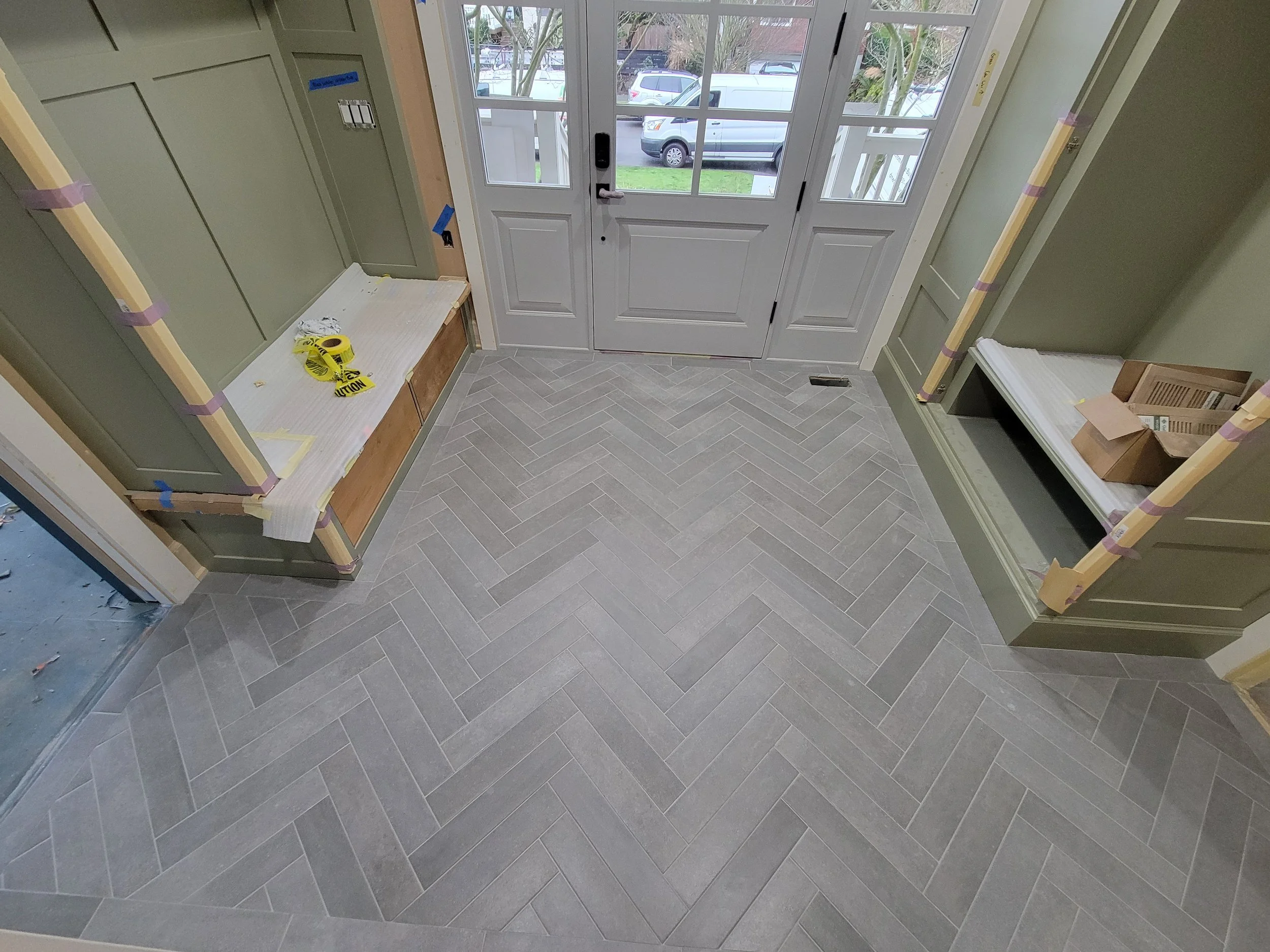 Entryway with herringbone tile flooring, green walls, and double glass doors leading outside. Shelves are being installed, with tools and materials on the surfaces, indicating ongoing construction or renovation.