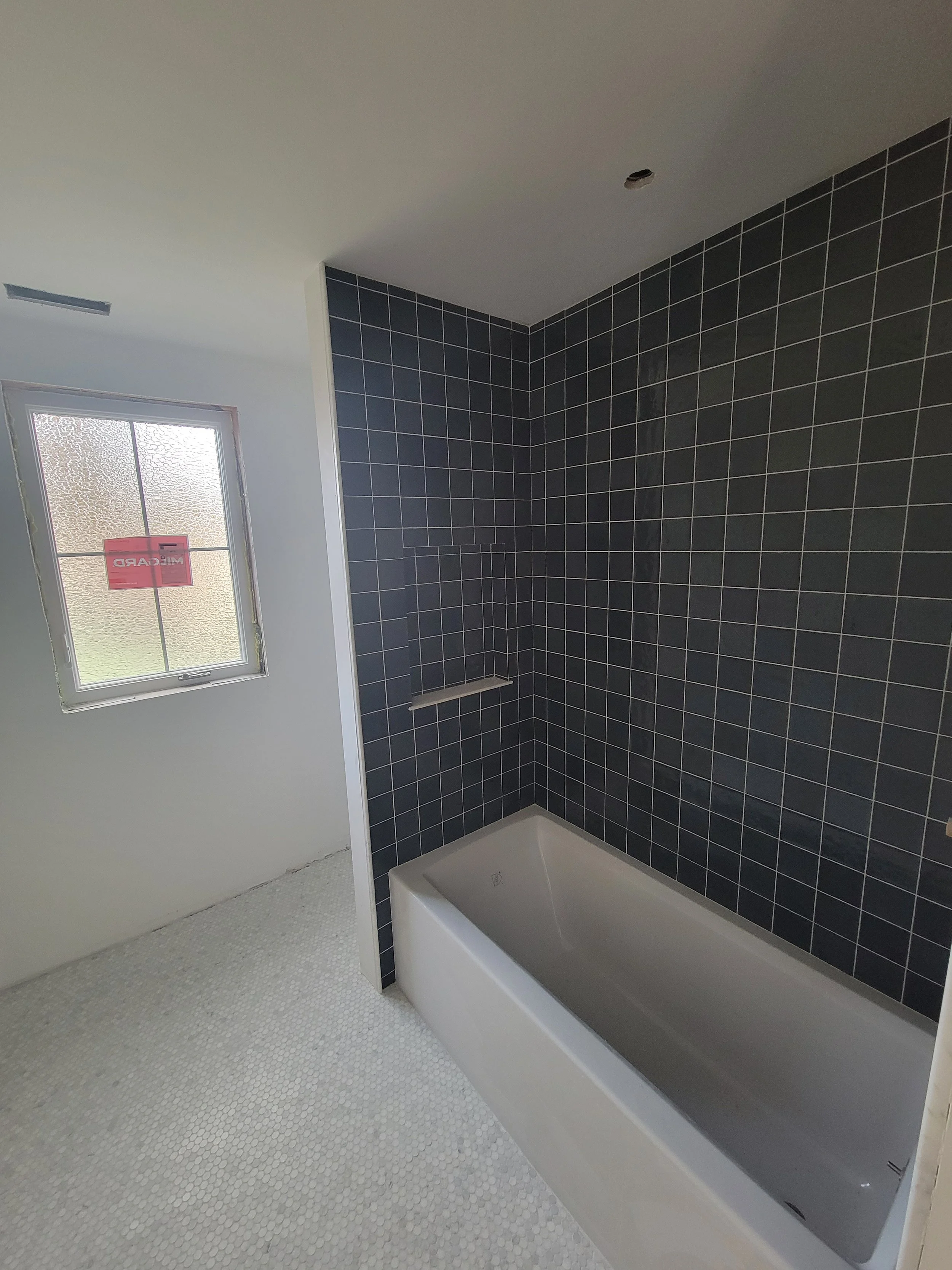 Bathroom with dark tiled walls and white bathtub, window with frosted glass, and white pebble-textured floor.