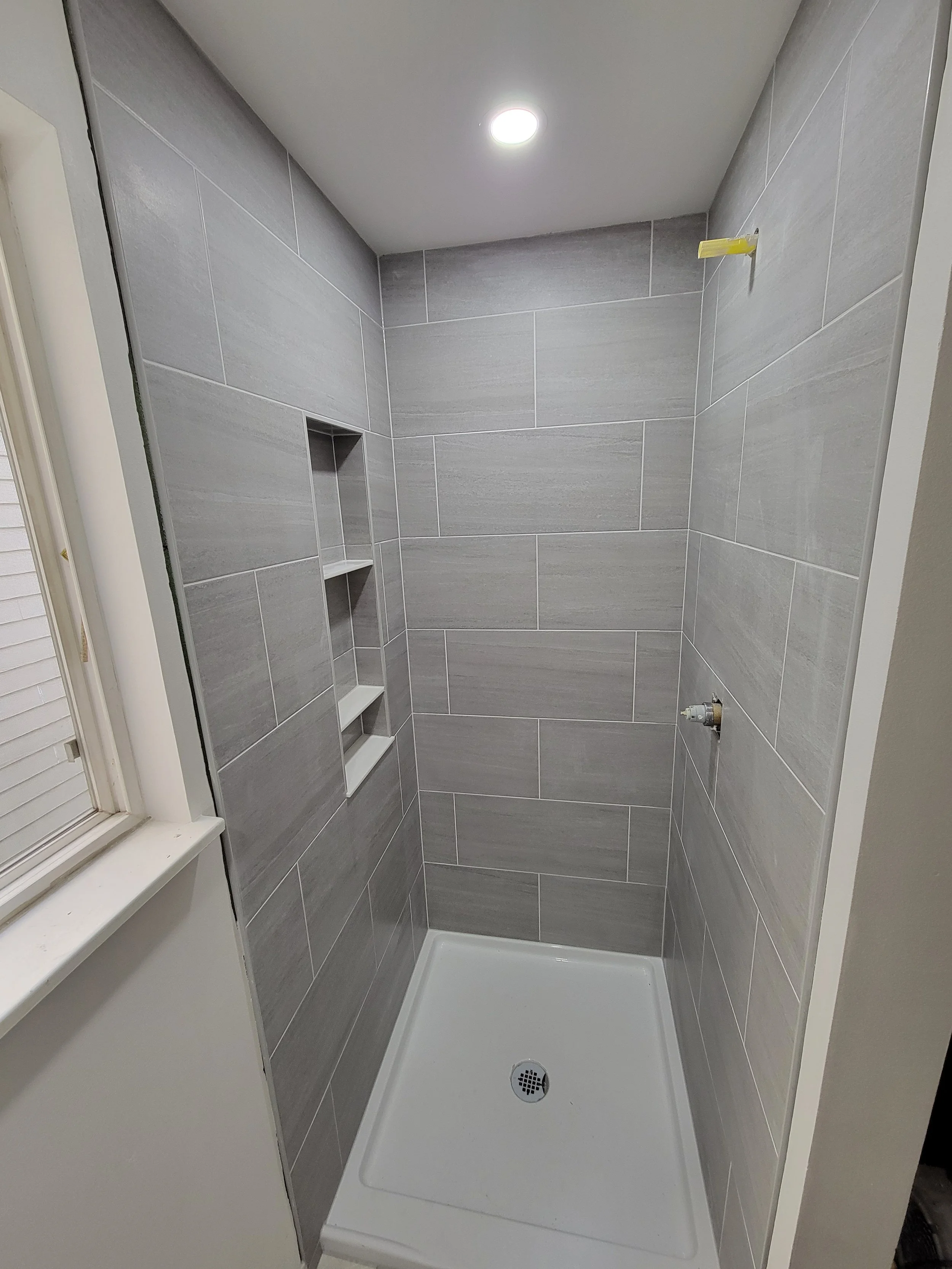 Empty shower stall with gray tiled walls, built-in shelves, a drain on the white shower base, and a ceiling light.